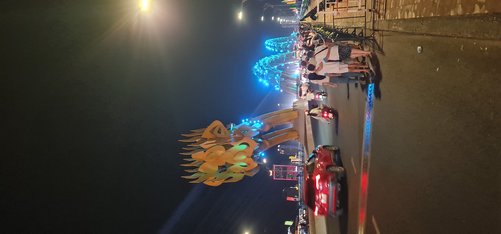 This night photograph captures a vibrant scene at the Dragon Bridge in Đà Nẵng, Vietnam. The iconic golden dragon head of the bridge is prominently featured in the foreground, illuminated by warm lights that highlight its intricate scale-like design and details. Its eyes appear to glow green, and parts of its body structure are lit with bright blue lights, extending along the bridge arches into the distance.
On the right side of the image, a sidewalk runs parallel to the bridge, where a group of people are gathered. They are casually dressed, with some standing and others leaning against a railing, appearing to observe the illuminated bridge. The street next to the sidewalk shows ongoing traffic, with a distinct red car in motion in the foreground, its taillights glowing. Several motorbikes are also visible on the road, some blurred by movement.
The time of day is clearly night, with the dark sky contrasting sharply with the numerous artificial lights. Bright streetlights create lens flares in the upper left and right portions of the image, adding to the nighttime ambiance. In the background, beyond the bridge, distant city lights and the faint outlines of buildings can be seen, indicating an urban setting. The overall scene suggests a lively atmosphere, likely a popular tourist spot. There is no discernible text visible in the image.
