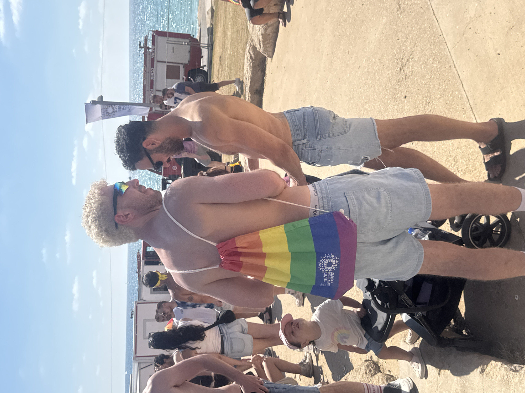 The image depicts an outdoor scene, likely a public event, on a sunny day. In the foreground, two shirtless men are standing and facing each other. The man on the left has blonde, curly hair and is wearing sunglasses. He has a rainbow-colored drawstring bag slung over his shoulder. The man on the right has dark, curly hair and is also wearing sunglasses. Both men are wearing light blue denim shorts.
To the left of the men, a young child wearing a light-colored t-shirt and a pink baseball cap is looking upwards. A stroller is positioned next to the child.
In the background, several other people are visible, some standing and some sitting. Behind the people, there are white structures that appear to be temporary booths or buildings. Further in the background, a red fire truck with the word "שירות" (service) and "כבאות" (firefighting) written on it in Hebrew is parked. Beyond the fire truck, the blue sea is visible. The overall environment suggests a coastal location in Tel-Aviv, Israel, possibly during a Pride event given the rainbow bag. The ground is paved and sandy in some areas.