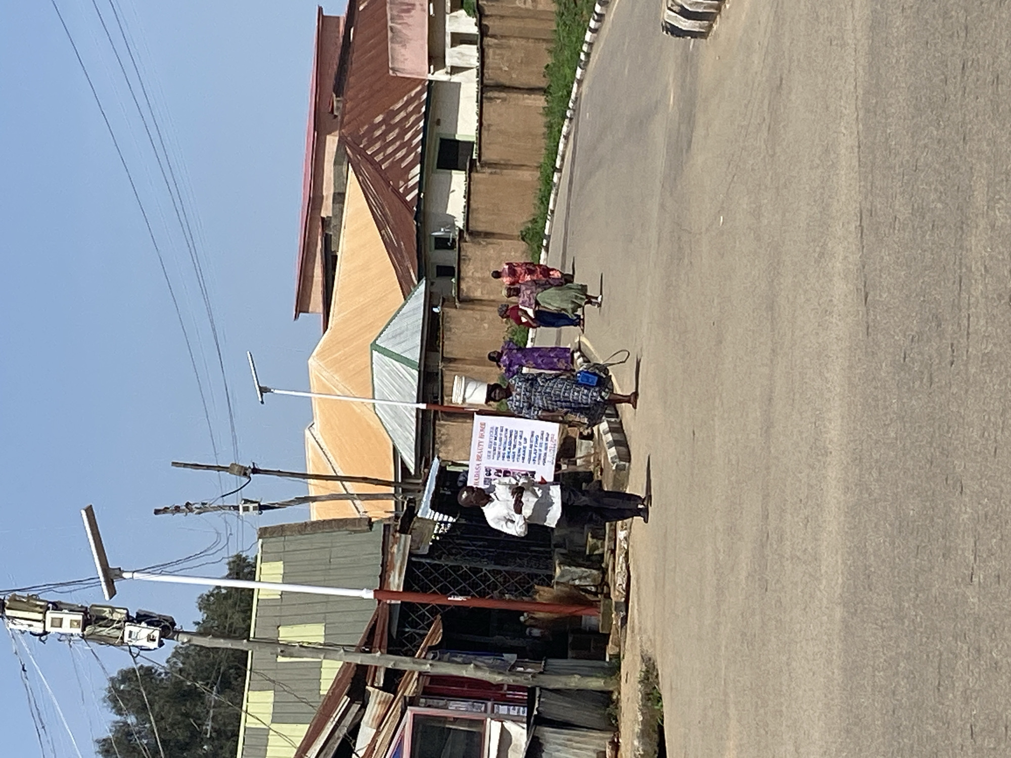 The image captures a street scene in Jos, Nigeria, on a clear, sunny day. In the foreground, a paved road curves to the right, with a curb on the right side marked with black and white stripes. A man in a white shirt and dark pants walks on the road, holding a white sheet of paper. To his right, a woman in a patterned dress carries a large white bucket on her head.
Along the left side of the road, a row of buildings with corrugated metal roofs and varying facade materials line the street. Some appear to be small businesses or shops. A prominent lamppost with several electrical boxes is situated near the buildings. In the midground, several other individuals, mostly women in colorful clothing, are walking further down the road.
The background consists of more buildings with diverse roofing materials, including corrugated metal and tiled roofs, as well as a concrete wall running along the right side of the street. Overhead, power lines crisscross the clear blue sky. The overall environment suggests a typical urban or semi-urban setting in Jos, characterized by a mix of commercial and residential structures, busy streets, and everyday life.