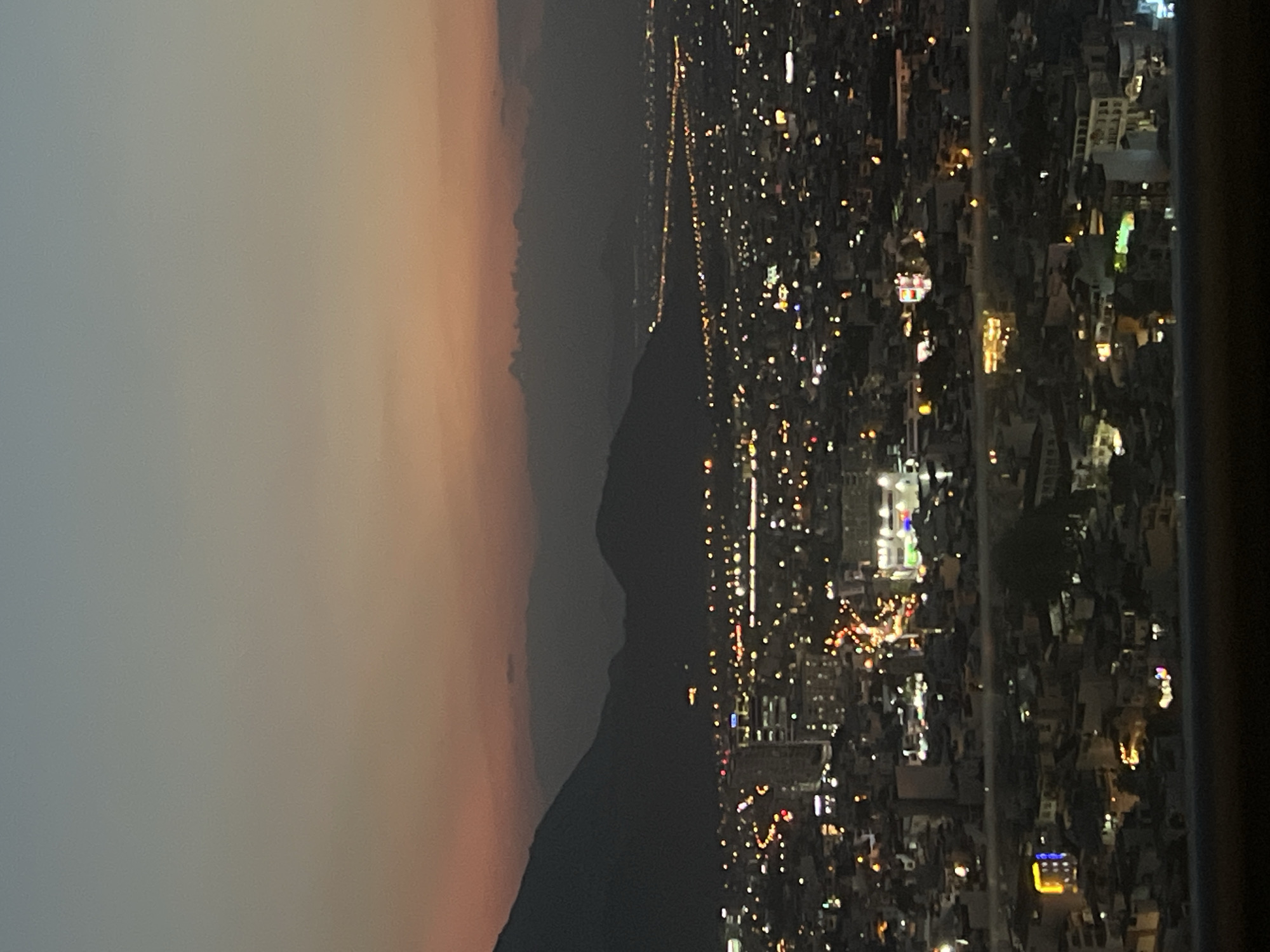 This is a nighttime, high-angle, medium shot of a city with mountains in the background. The sky is a gradient of dusky orange and light blue, transitioning to a darker gray near the mountains. The city is illuminated by a multitude of lights from buildings and streetlights, creating a vibrant, sprawling landscape. In the foreground, there are residential buildings with some illuminated windows and colored lights. Further into the city, taller buildings and more densely packed lights are visible. A long, winding line of lights suggests a major road or highway extending into the distance. The mountains are dark silhouettes against the twilight sky. No people are visible. The overall impression is of a bustling urban environment at dusk.
