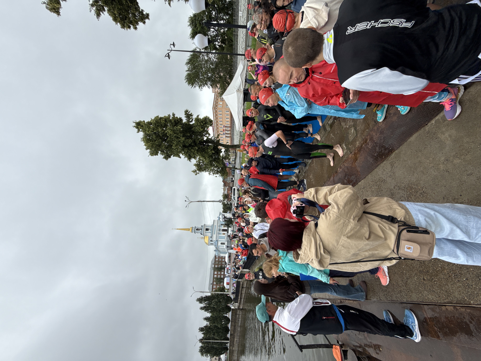 The image depicts a gathering of people, many wearing red swim caps, standing by a body of water in Votkinsk, Russia. The backdrop features a light blue church with a golden dome and spire, characteristic of Russian Orthodox architecture. Several trees are visible, and the sky is overcast, suggesting a cloudy day. Some individuals are dressed in wetsuits, indicating a possible swimming event or competition. A woman in the foreground is holding a camera, appearing to take photos of the assembled group. People are standing on a paved area next to a railing overlooking the water. The overall scene suggests an outdoor event or organized activity taking place near a waterfront in Votkinsk.