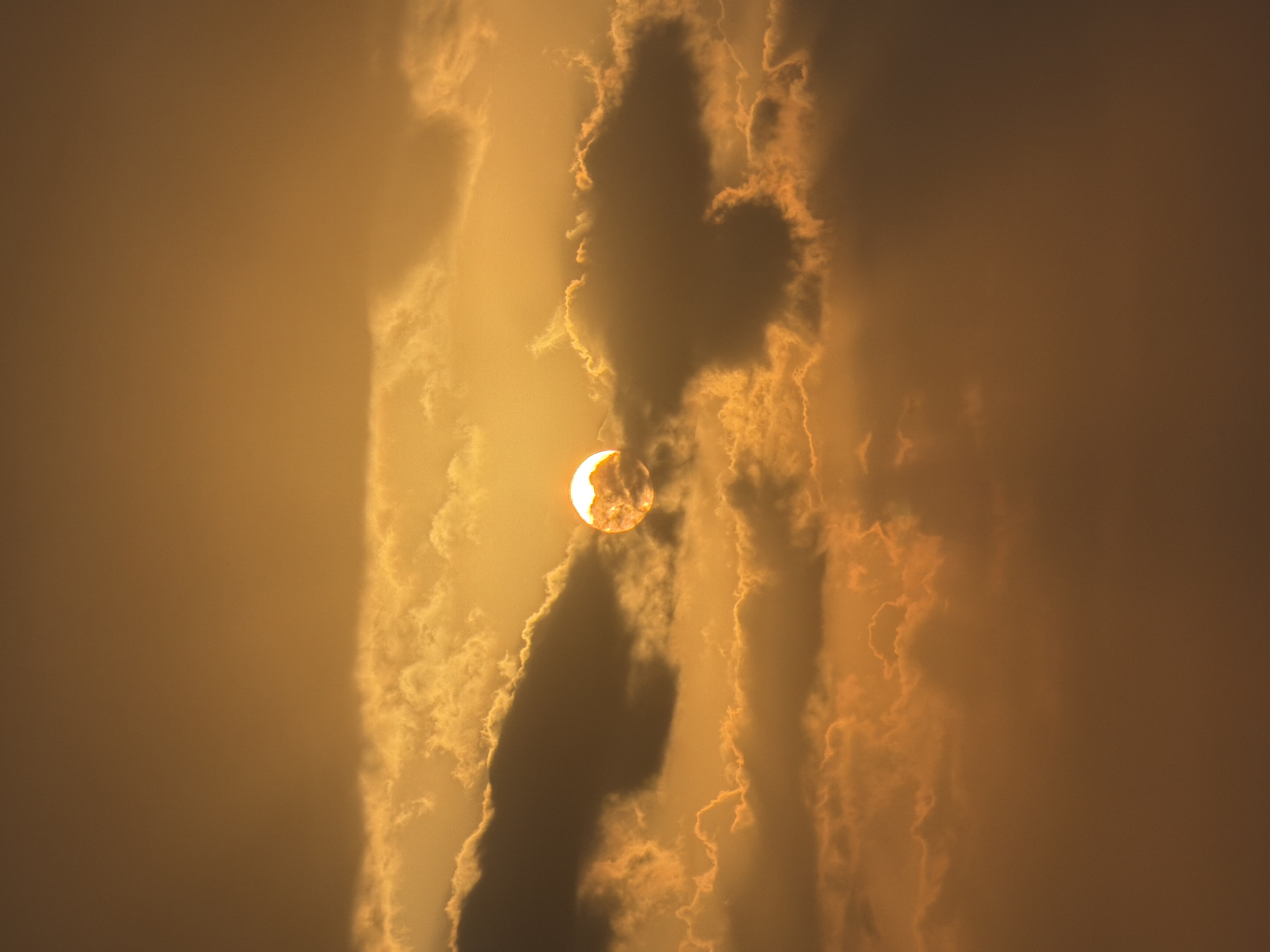 A sun appears partially obscured by clouds. The sun is round with a bright orange glow. Dark gray clouds surround the sun, with lighter orange clouds in the background. The sky is a gradient of orange hues.