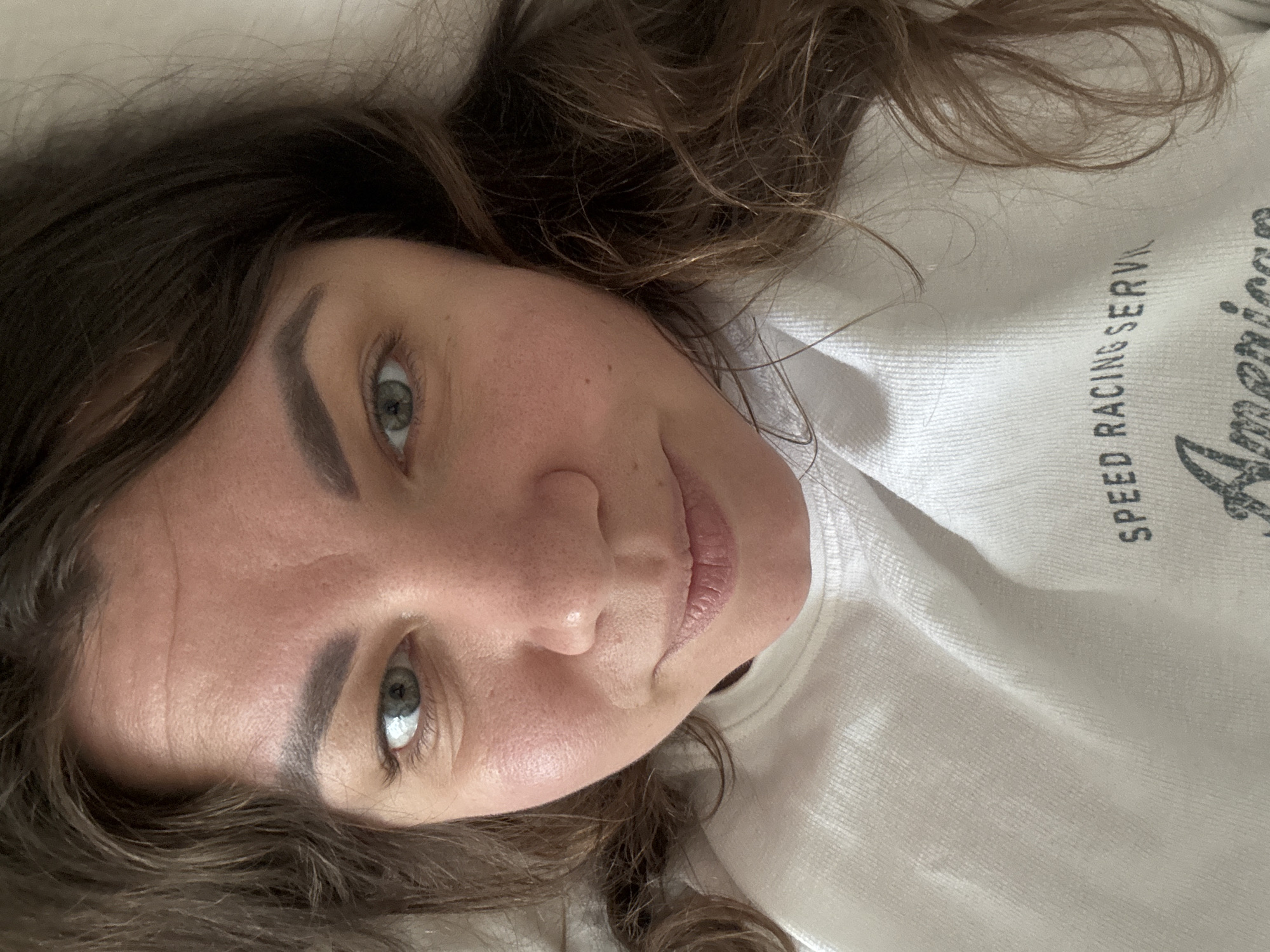 A woman with dark brown, wavy hair is lying down and looking directly at the camera. She is wearing a white sweatshirt with text that appears to say "SPEED RACING SERVICE" and "American." The lighting is soft and diffused, suggesting an indoor setting. There is no indication of the location being in Höhenkirchen-Siegertsbrunn, Germany.