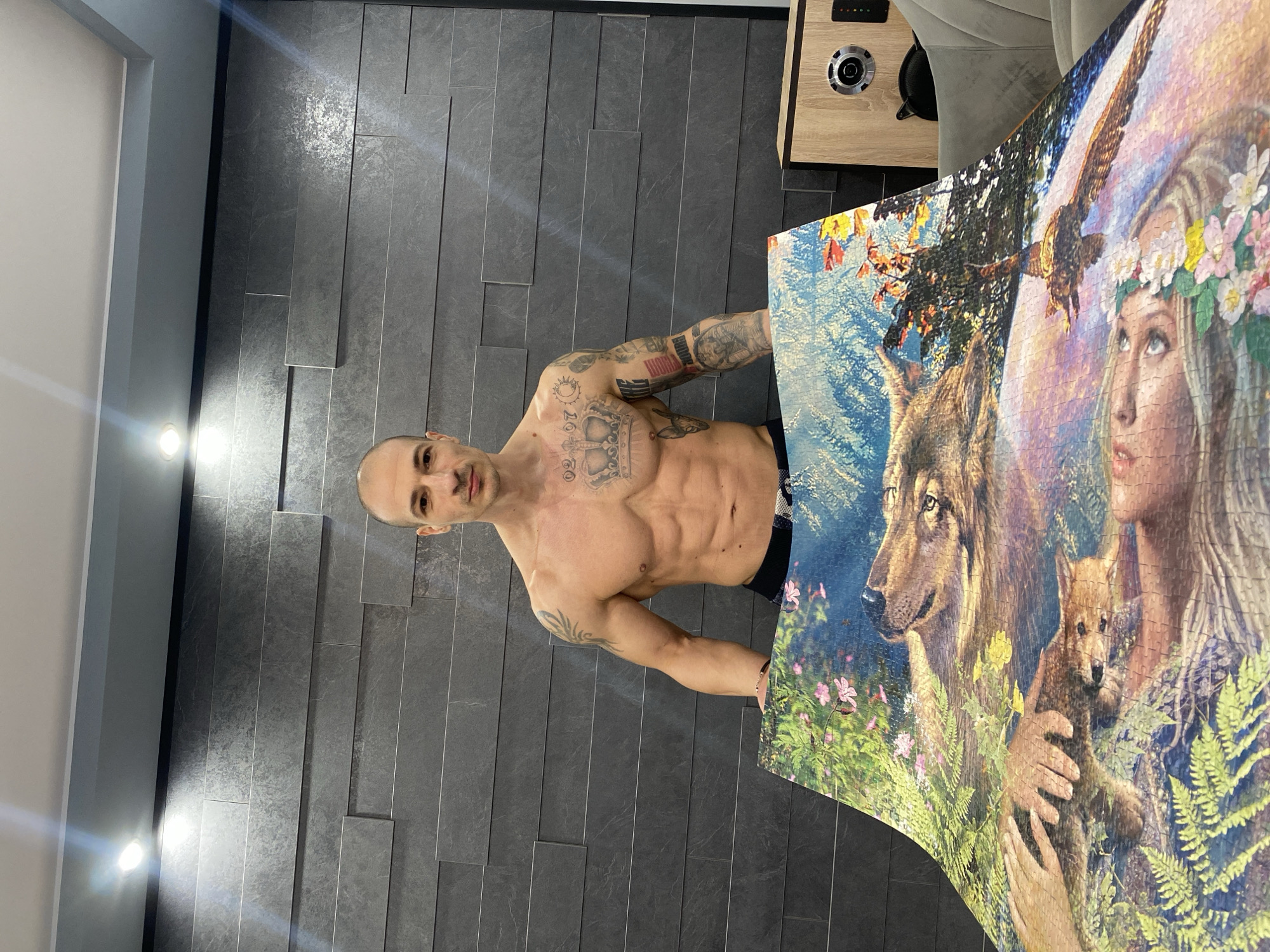 The image shows a shirtless man standing in a room with a gray tiled wall, holding a large jigsaw puzzle. The puzzle depicts a woman with a flower crown, an eagle, a wolf, and a wolf cub. To the right of the man is a wooden cabinet and a gray chair. The location is Pleven, Bulgaria, based on the user's prompt.