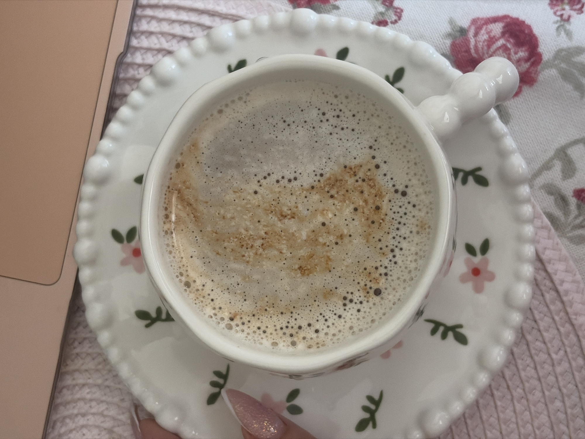 A close-up overhead shot shows a cup of coffee resting on a saucer, positioned on a surface that includes a floral-patterned fabric and part of a laptop. The coffee is light brown with a foamy surface. The saucer is white with a beaded edge and a floral pattern of small pink flowers with green leaves. To the left of the saucer, part of a laptop is visible with a peach-colored casing. The background consists of two different fabric textures; one is a pale pink woven material, and the other has a white floral print. A person's fingernail is visible in the bottom center, painted with glitter polish. There is no indication of the location being Saint Petersburg, Russia.