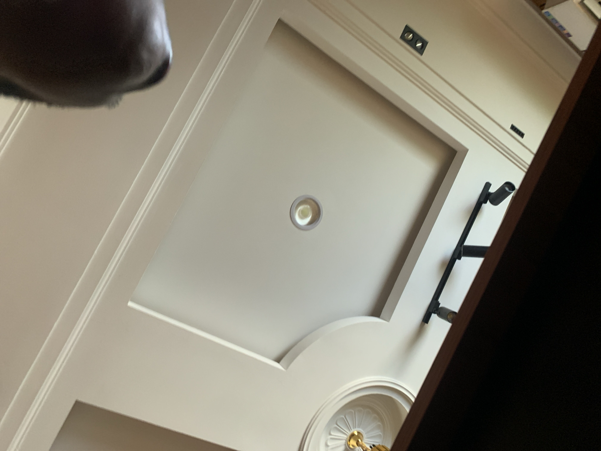 The image captures a low-angle view of a ceiling with recessed lighting and decorative molding. In the upper right portion of the image, a person's head and neck are partially visible, suggesting they are looking upwards. The ceiling features a central recessed square with a light fixture, surrounded by layered molding. To the left, a decorative circular element with golden accents is visible. A track lighting system with two adjustable spotlights is mounted on a lower part of the ceiling, and further to the right, two rectangular light fixtures are embedded. The overall impression is of an interior space with modern and classic design elements. The location in Gura Top, Nigeria, is not directly discernible from the visual elements of the ceiling.