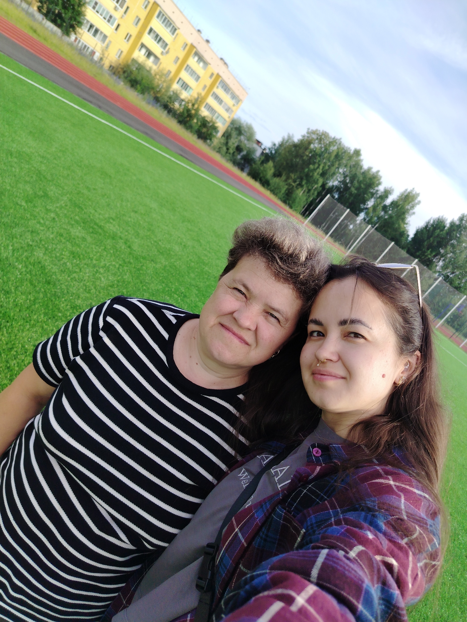 This image is a selfie taken outdoors at a sports field in Noviy, Russia. Two people are visible. On the left is an older woman with short, graying hair, wearing a black and white horizontally striped shirt. To her right is a younger woman with long, dark brown hair, wearing a purple and blue plaid shirt over a grey t-shirt. She has a nose ring and earrings. They are both smiling and looking at the camera. The background consists of a bright green artificial turf field, a red running track, a yellow apartment building, trees, and a cloudy sky.