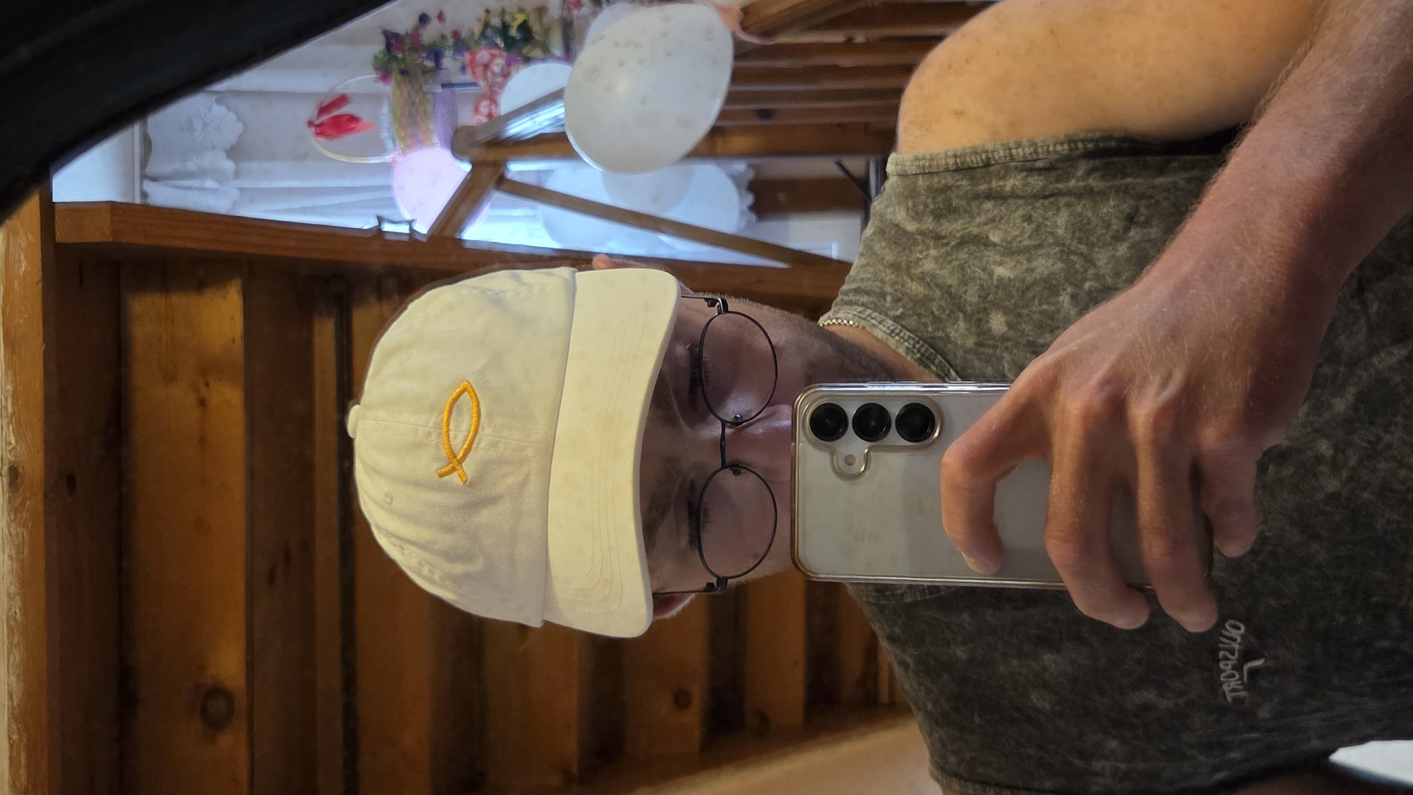 A mirrored selfie captures a person wearing a white baseball cap with a golden fish symbol and round glasses. The person is holding a smartphone. Reflected in the mirror are wooden beams, white balloons, and decorative floral arrangements, all indicating an indoor setting, possibly a residence in Bania, Bulgaria during a celebratory event.