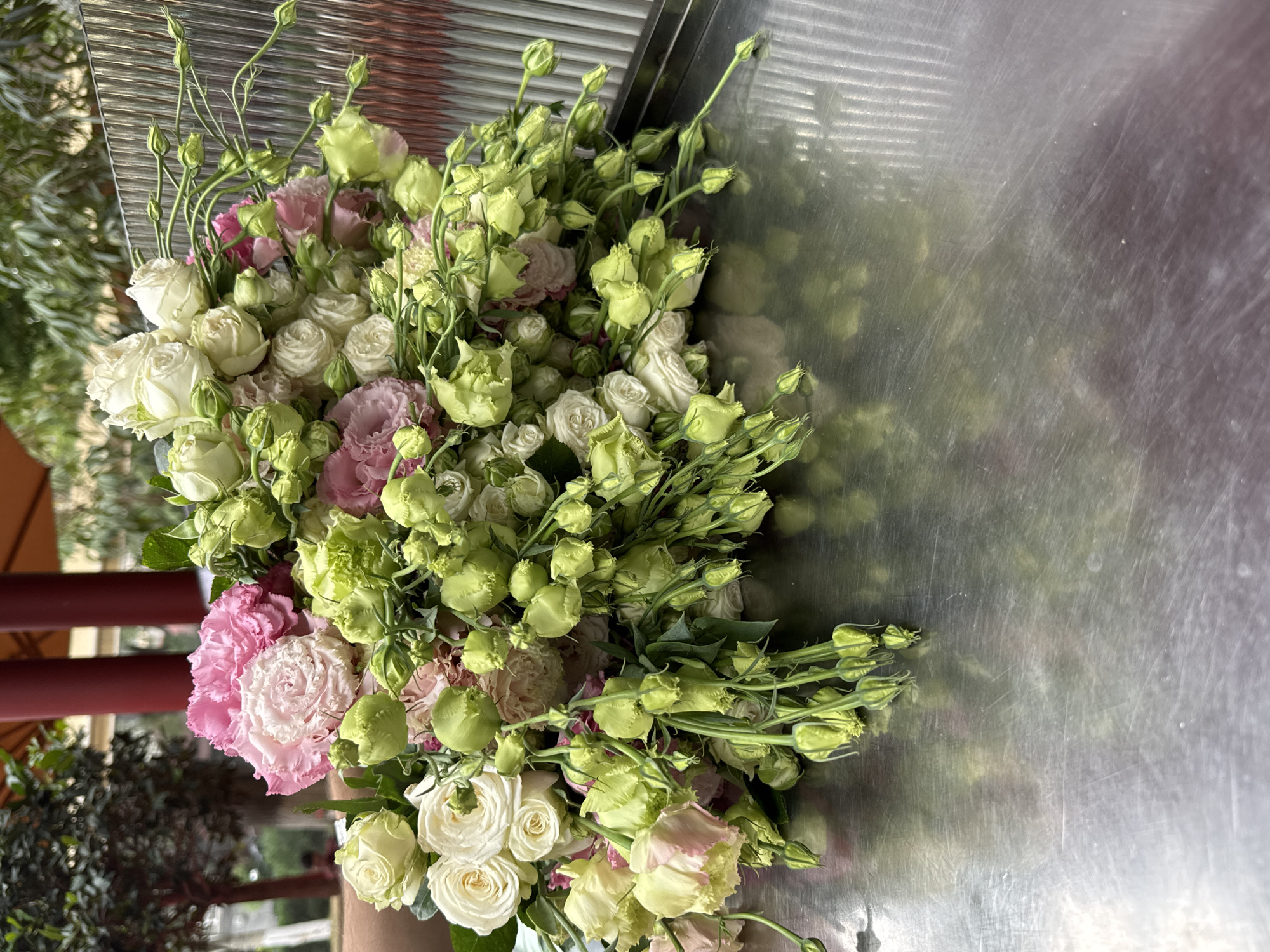 A large bouquet of white and pink roses, mixed with green buds and stems, rests on a reflective metal surface, likely a table. The bouquet is lush and full, with the flowers cascading downwards. In the background, out of focus, are red pillars and greenery, suggesting an outdoor or semi-outdoor setting. The metal surface reflects the bouquet and the background, creating a distorted and shimmering effect. The image is well-lit, with natural light highlighting the textures and colors of the flowers. There is no indication of the city of Yerevan, Armenia, within the image itself.