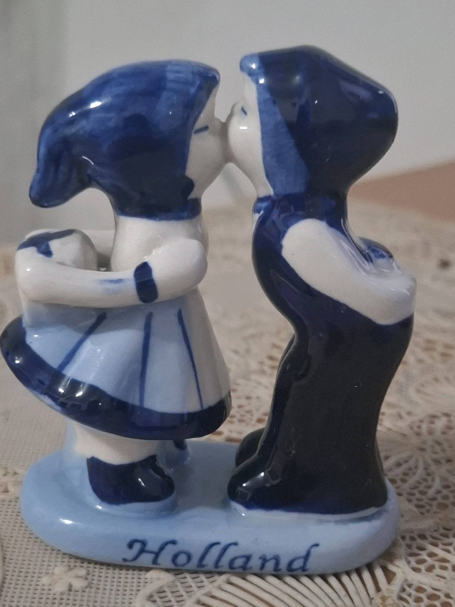 A ceramic figurine of two children in traditional Dutch attire, one dressed in blue and white with a light blue skirt, and the other in a dark blue outfit, are depicted in a kiss. The base of the figurine is light blue and inscribed with the word "Holland" in blue lettering. The figurine is placed on a white lace tablecloth, with a blurred background of a light-colored wall and a brown surface. The lighting in the scene is soft and diffused. The image's context of Almaty, Kazakhstan is not discernible from the visual information provided.