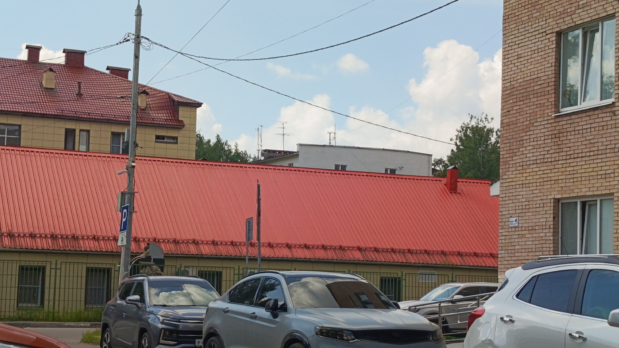 The image shows a street scene in Zvenigorod, Russia, dominated by a large building with a vibrant red corrugated metal roof. To the left of the red-roofed building, a lighter colored building with a red tiled roof and dormer windows is visible. In the foreground, several cars are parked or driving, including a silver SUV and a white SUV. A light pole with a blue parking sign is positioned near the center of the image. The sky is partly cloudy, with a clear blue expanse and fluffy white clouds. Trees are visible in the background, interspersed with other buildings. The overall impression is that of an urban or suburban environment on a sunny day.