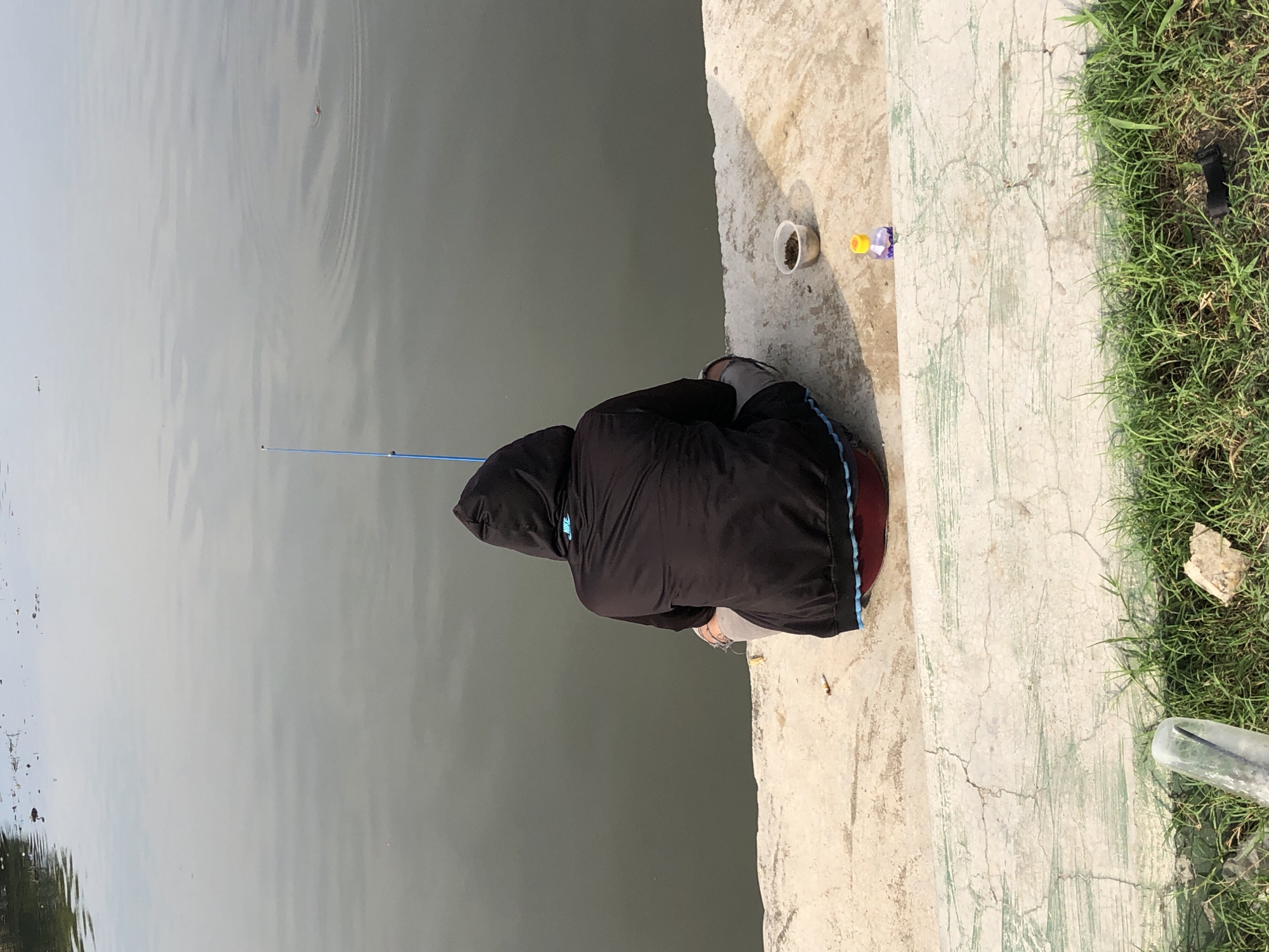 A person is sitting on the edge of a concrete embankment, facing a body of water. They are wearing a black hooded jacket and gray pants, and are holding a blue fishing rod. A small container of bait is placed next to them. The water appears calm with some ripples. The embankment is cracked and weathered, with patches of green paint. In the foreground, there is green grass and some debris. This scene is likely in Lhokseumawe, Indonesia, possibly a canal or riverbank commonly used for fishing.