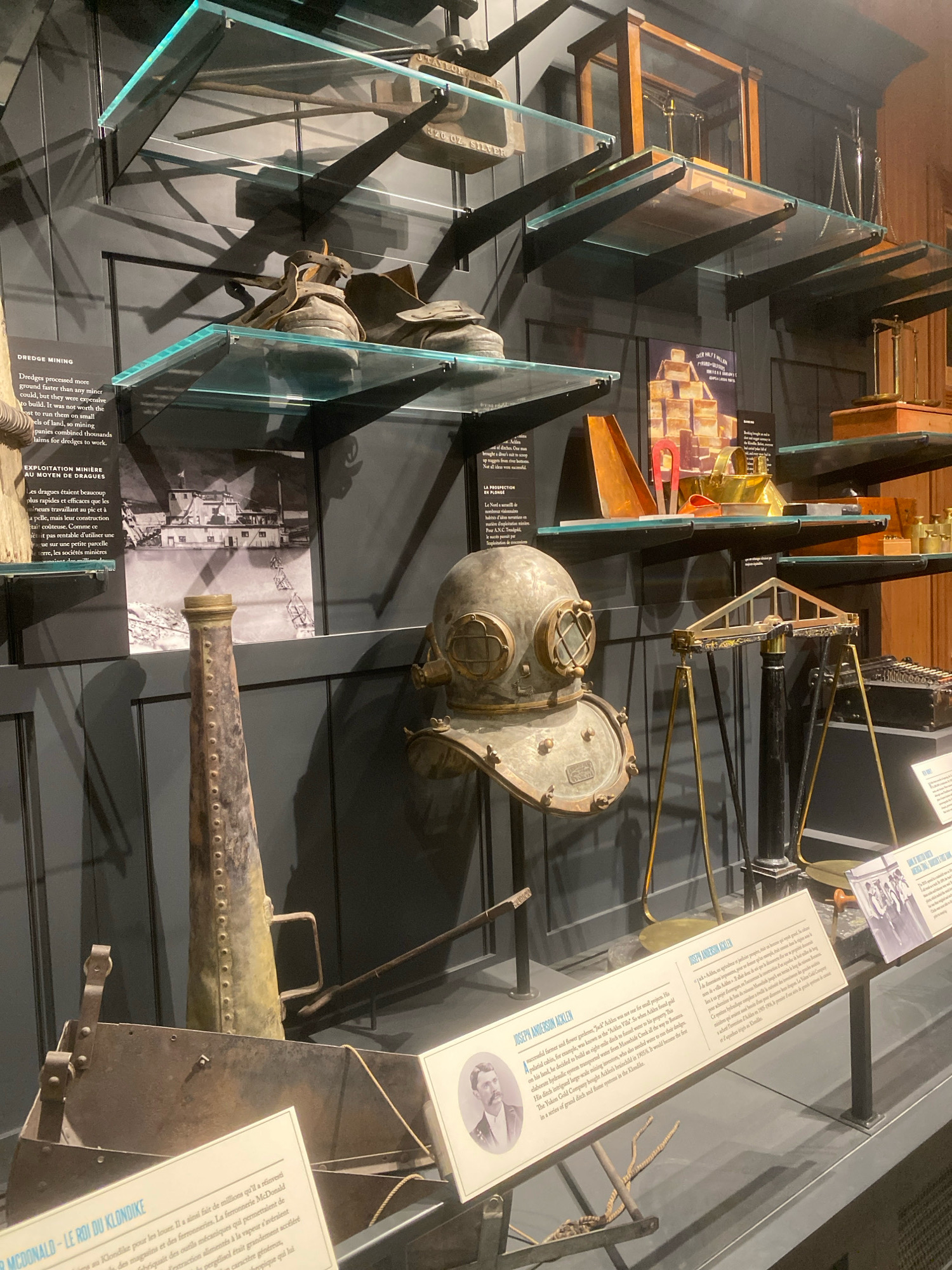 The image displays an exhibit at the Dawson City Museum in Dawson City, Canada, showcasing historical mining equipment and artifacts. In the foreground, a large, weathered diving helmet is prominently featured, positioned to the left of a brass balance scale. To the left of the diving helmet is a tall, conical metal object with rivets, and in front of these items is a rusty metal sluice box.
The background features shelves displaying various items. The top shelf holds what appear to be old tools, possibly for mining. The shelf below that has a pair of worn boots or shoes. Further down, a framed black and white photograph depicts a large dredge boat on a body of water, with text in English and French to its left and right. Another shelf displays a collection of what appear to be gold bars or ingots, alongside other metal objects. The overall environment is a museum exhibit with a dark, gray wall as the backdrop, contrasted by illuminated display shelves. Informational placards with text and images are visible at the bottom of the display.