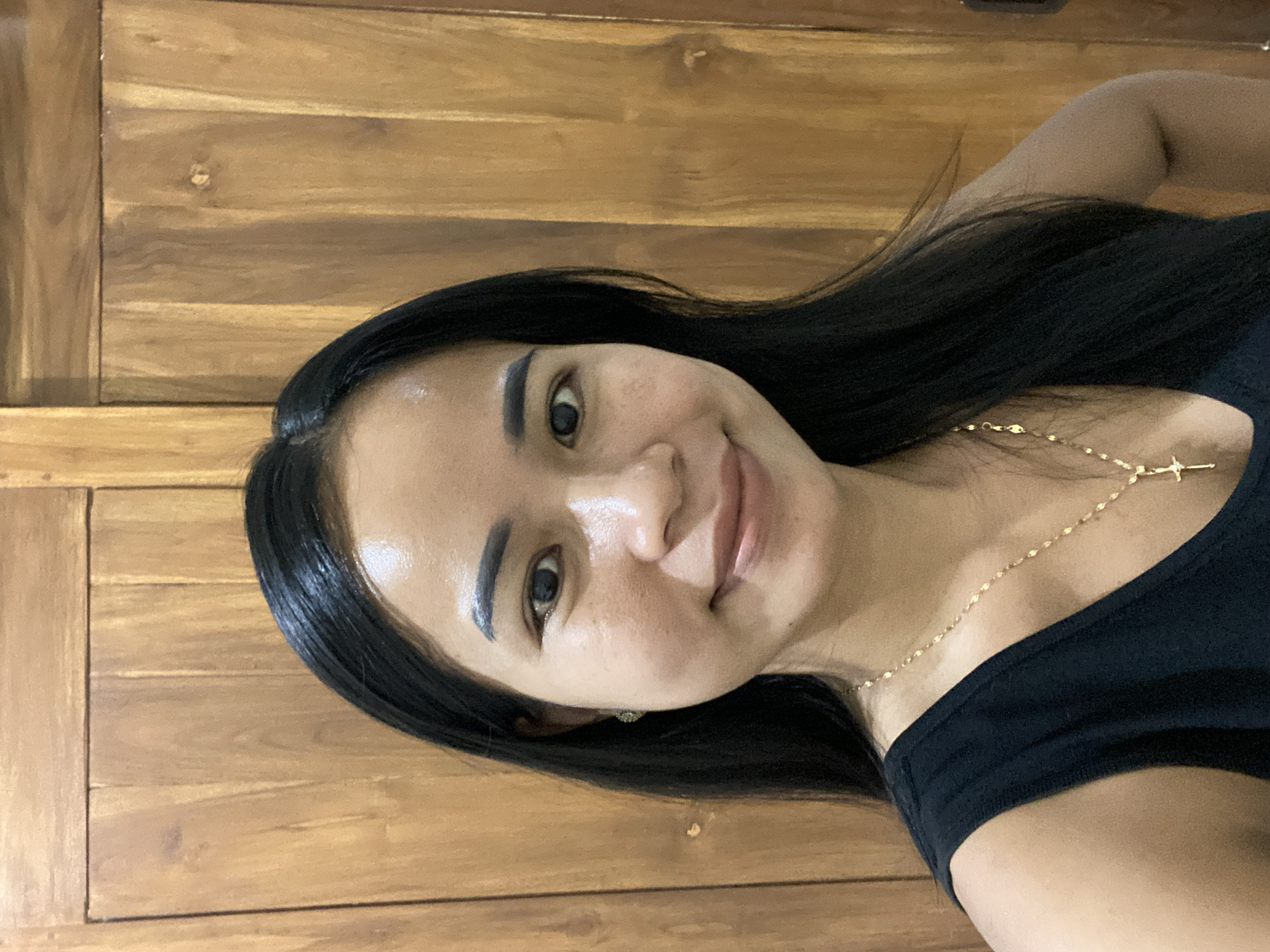 A selfie of an Asian woman is set against a light brown wooden panel background. The woman has long, straight, black hair and is wearing a black shirt. A gold chain necklace with a cross pendant is visible on her chest. She has smooth skin, dark eyebrows, and appears to be smiling slightly. The location and city in North Kuta, Indonesia, is not deducible from the provided picture.