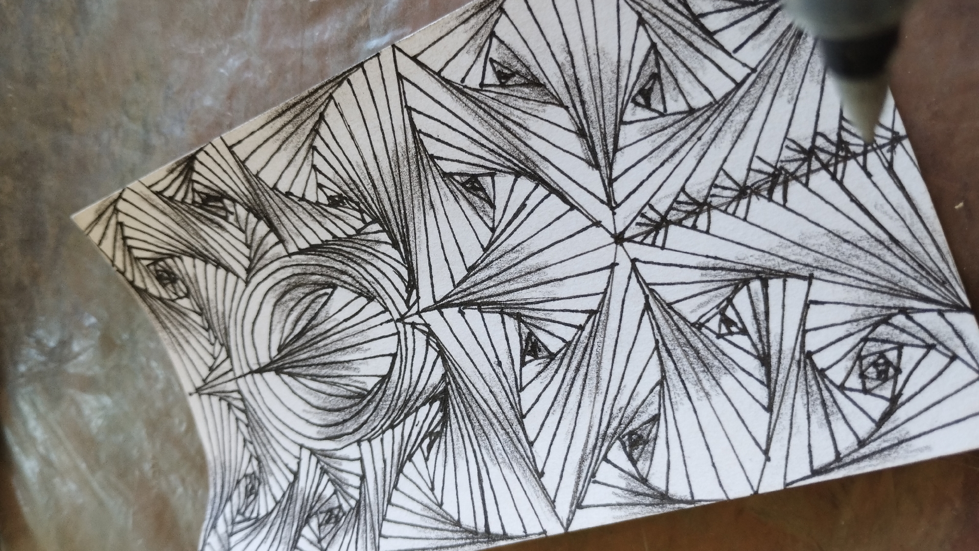 This is a close-up, eye-level, indoor shot of a hand-drawn geometric pattern on a piece of white paper. The drawing features intricate black lines that create a sense of depth and dimension, with many converging lines forming triangular shapes and spirals. The paper is slightly curved, and it rests on a textured, brown surface. In the bottom right corner, the tip of a pen or marker is visible, suggesting that the drawing is in progress. The background is blurred and appears to be a gray, reflective surface. There are no people or any identifiable elements of a city.