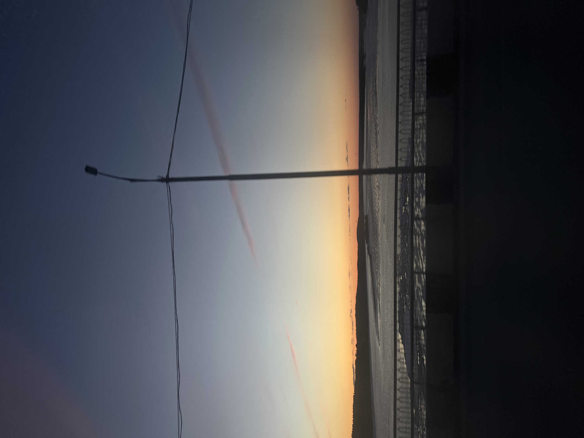 The image shows a landscape view at dusk in what appears to be Usolye, Russia, based on the location context. The sky transitions from blue to orange near the horizon, with faint streaks of clouds visible. In the foreground, there's a lamppost with wires extending across the frame. Adjacent to the sky, what is perceived as a road or pathway can be seen, followed by buildings in the distance. The right part of the image has a dark area, obstructing a complete view of the environment.