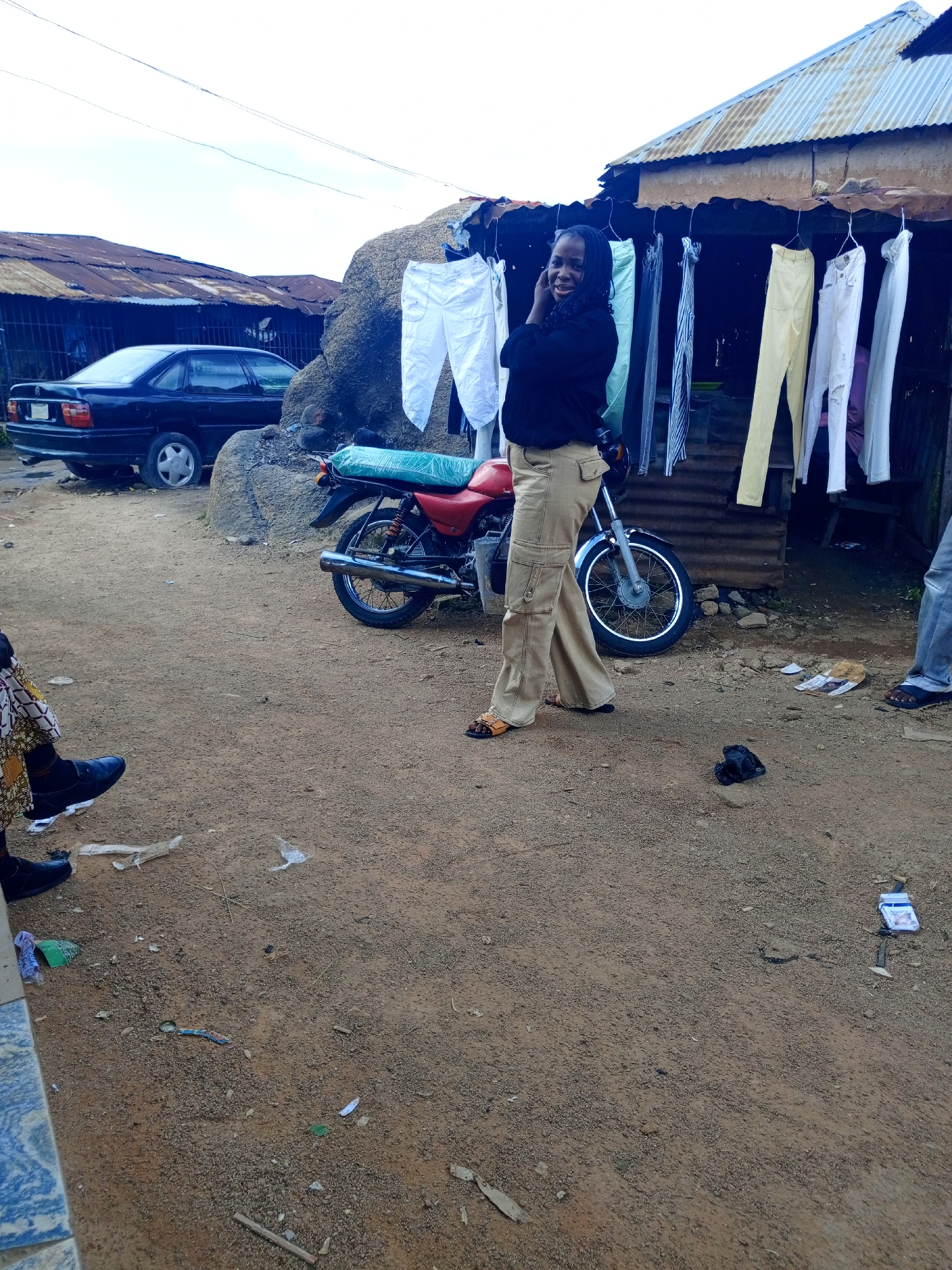 In Jos, Nigeria, a young woman stands outside a small roadside stall. The stall has a corrugated metal roof and displays a variety of clothing items, including pants and shirts, hanging on hangers. The ground is dirt, with scattered debris. To the left of the stall, a black sedan is parked. A red motorcycle with a green seat cover is parked in front of the stall, and a large rock formation is visible behind it. The woman, wearing a black top and tan pants, is looking towards the camera and appears to be smiling. Another person's legs are visible in the lower left of the frame, wearing a patterned skirt and black shoes. In the lower right, another person's legs are visible, wearing blue jeans and sandals. The overall scene depicts a typical street view in a Nigerian city, with elements of commerce and everyday life.