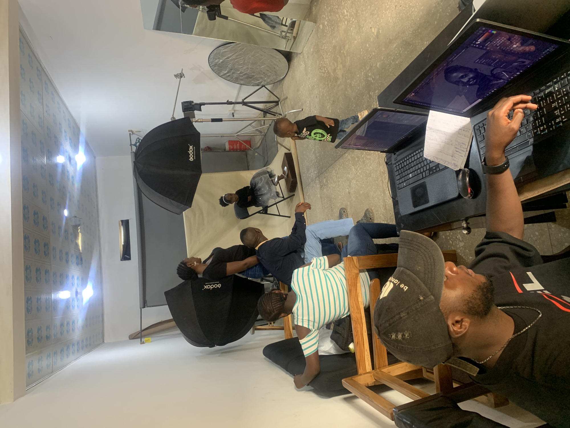 The image shows a photography studio. There are five people in the studio. One person is seated at a desk working on a laptop, two people are seated on chairs being photographed, and one person is standing and observing the models. There is also a child standing. The studio contains photography equipment, including softboxes and a reflector. The background is a white sheet. The studio is located in Gura Top, Nigeria.