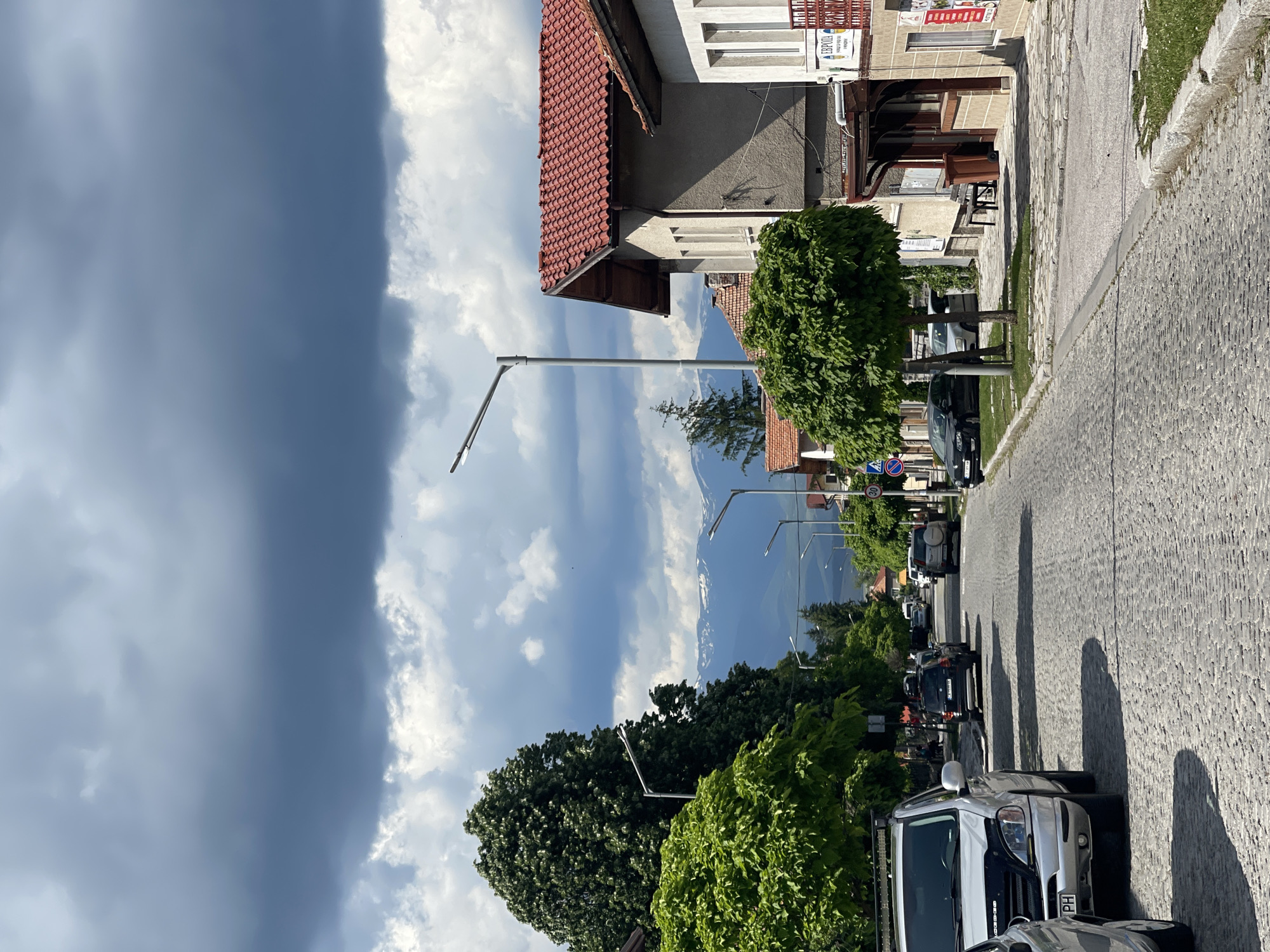 The image depicts a street scene in what appears to be a town or village, with snow-capped mountains visible in the distance under a dramatic, cloudy sky. The street is paved with cobblestones and lined with buildings and trees. Several cars are parked on the side of the street, and some appear to be in motion. On the right side of the street, there are commercial buildings with tiled roofs and storefronts. On the left side, there are residential buildings and mature trees. The overall atmosphere suggests a somewhat overcast day with a touch of rugged natural beauty from the mountains.