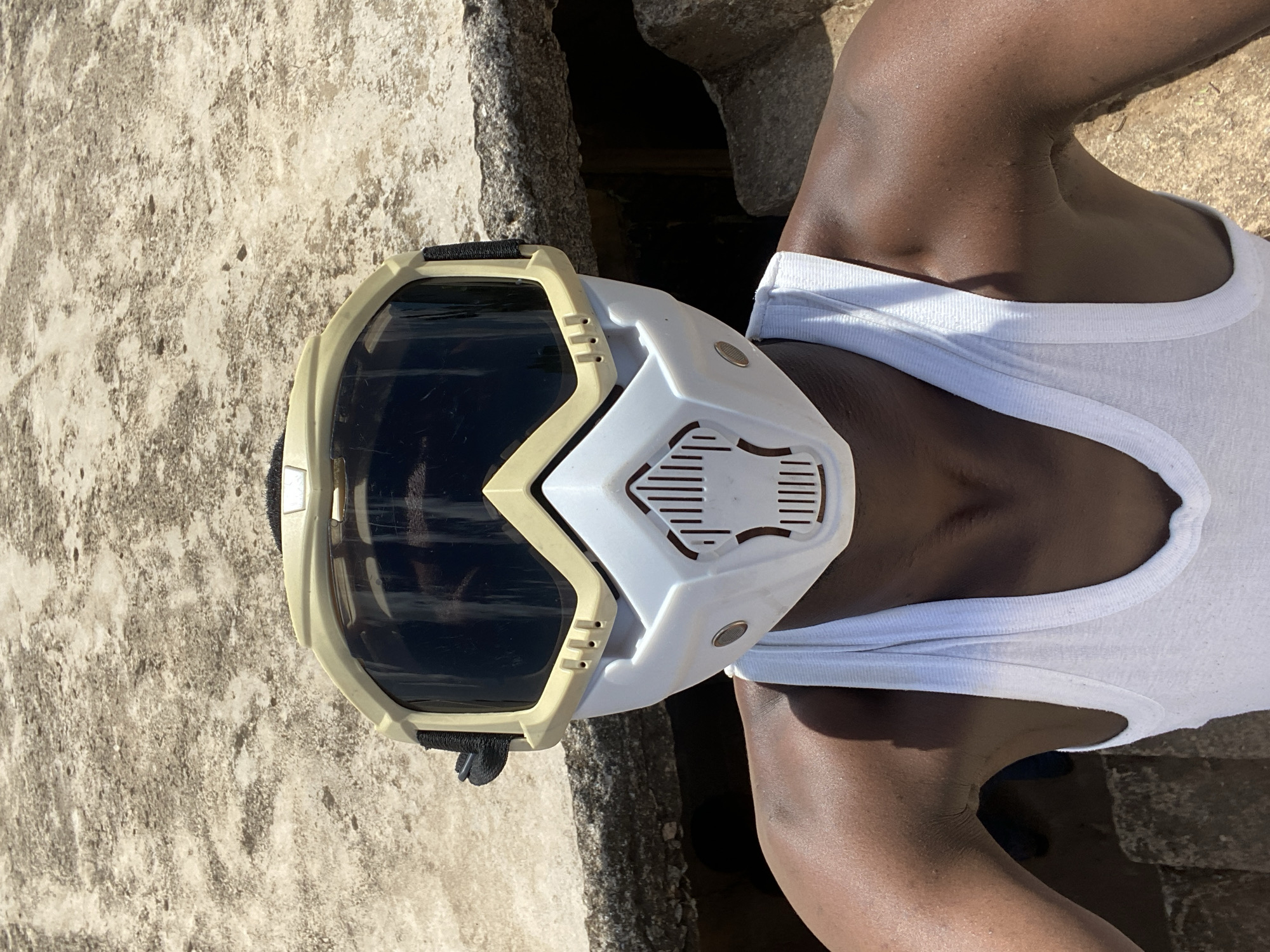 A person wearing a white tank top and a white and khaki full face mask is positioned in front of a weathered concrete structure. The person's skin tone is dark. The mask features dark lenses and ventilation slots on the lower portion. The concrete surface is rough and uneven, showing signs of aging. The location is Bukuru, Nigeria.