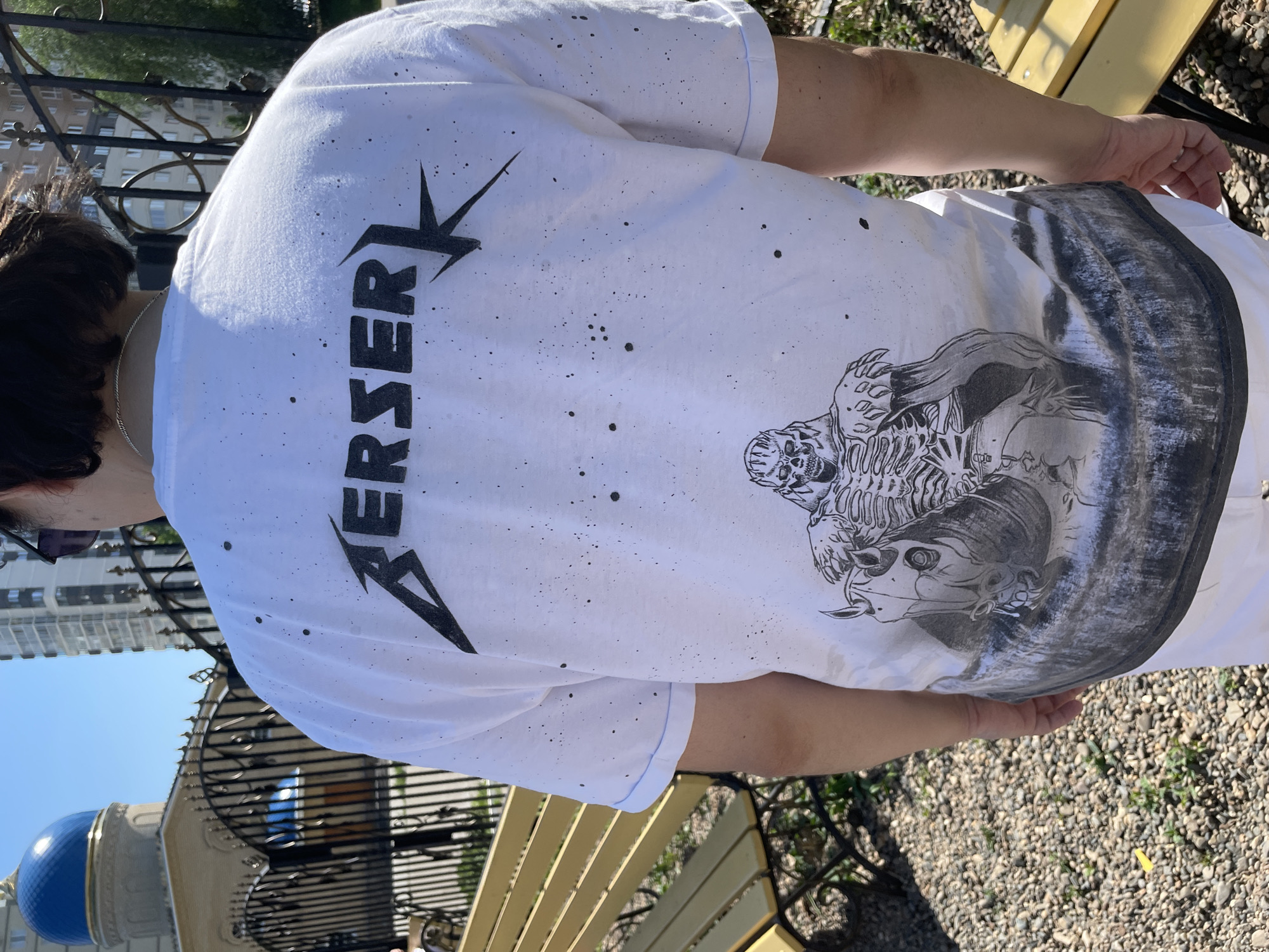 The image shows a person wearing a white T-shirt with "BERSERK" printed vertically in black on the back, along with a drawing of a skull-faced creature and a horse. The shirt is also marked with small black dots. The person is standing outdoors, with a yellow bench partially visible in the foreground. In the background, there is wrought iron fencing, a building with a blue dome, and other buildings, suggesting an urban setting. The image was taken in Izhevsk, Russia.