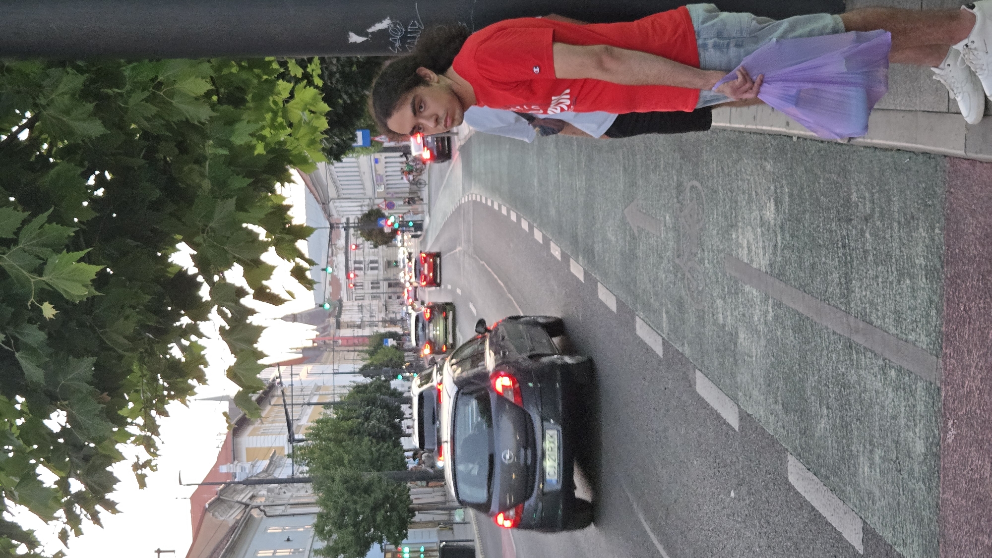 A vertical, eye-level shot shows a street scene in Cluj-Napoca, Romania, featuring traffic, pedestrians, and buildings. The foreground is dominated by a dark gray Opel car with its brake lights illuminated, driving on a multi-lane road with green and white markings. To the right, a young man with long, dark, curly hair and wearing a red t-shirt and denim shorts stands by a black pole, holding a translucent purple plastic bag in his right hand. Another person, wearing a light blue shirt and dark pants, stands slightly behind and to the left of the man in red. In the background, several other cars are visible, along with traffic lights showing red and green signals. Buildings with red-tiled roofs and light-colored facades line the street, and trees with green foliage are present. The sky appears overcast. The image captures a moment of urban life with a focus on traffic flow and pedestrians.