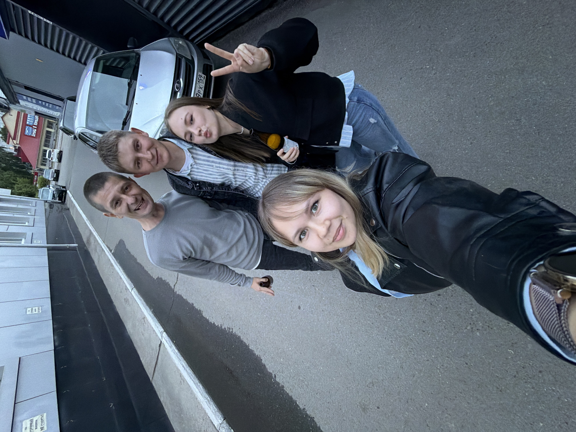 Four individuals, two men and two women, are captured in a high-angle selfie on an asphalt street in Chaikovsky, Russia. The street surface is damp in sections, with a light-colored curb visible on the left.
The woman in the foreground (bottom left) is taking the selfie, looking towards the camera with a slight smile. She has light brown hair and light eyes, wearing a black leather jacket over a light blue shirt. A gold-toned smartwatch with a mesh band is visible on her left wrist.
To her right, a man with short, dark hair smiles broadly, wearing a grey long-sleeved shirt and dark pants. He holds a dark-colored bottle in his right hand. Behind him, another man with short, light brown hair smiles, wearing a light-colored striped shirt under a dark denim jacket.
On the far right, a woman with long, dark brown hair poses with a "duck face" expression, displaying a peace sign with her left hand while holding a light-colored bottle in her right hand. She wears a black bomber jacket over a light blue and white striped shirt and ripped blue jeans.
In the background, a silver Lada Granta car with Russian license plate "9PX 159" (region code 159, indicating Perm Krai) is parked. Further down the street, additional vehicles are visible. Buildings line the street; one on the left features grey and black paneling with partially obscured Russian text including "НЕ СТАВИТЬ" (Do not park). A building with red roofs and a sign reading "ТЕАТР АРТЫ И КОВАНЫ" (Theater Arty and Kovany) is prominent in the distant background.