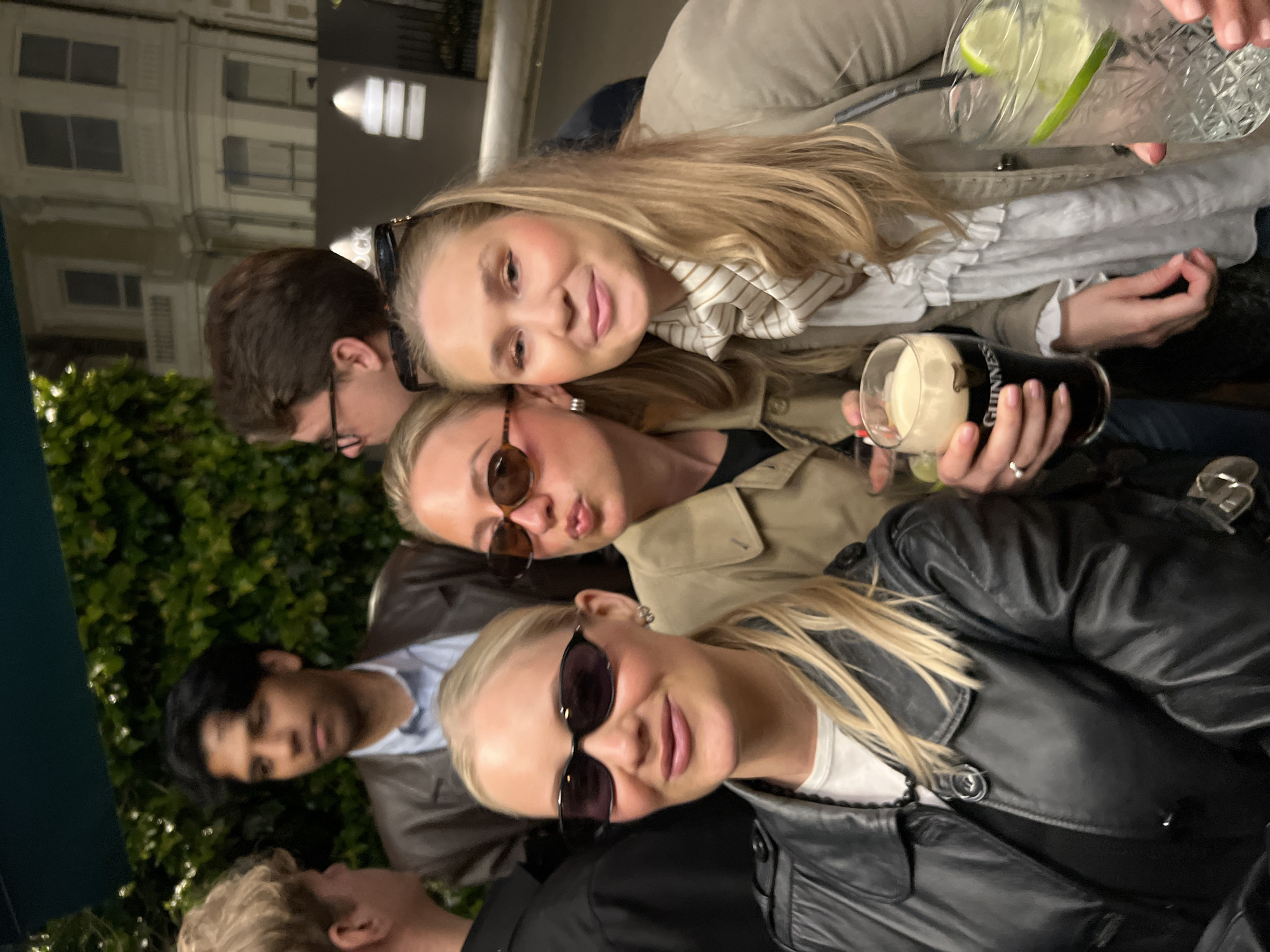 A group of people is posing for a photo outside a building in London, United Kingdom. Three women are in the foreground; all have fair complexions and blonde hair. The woman at the bottom is wearing a black leather jacket and sunglasses. The woman in the middle is also wearing sunglasses and a tan jacket, and the woman on top is wearing a light beige coat. The woman in the middle is holding a pint of Guinness. The woman on top is holding a tall glass with liquid and lime. To the left of the group, two men are visible. One has dark hair and is wearing glasses. The other man has dark hair and is wearing a light blue shirt and brown jacket. Behind the group is a green hedge and a building with illuminated windows.