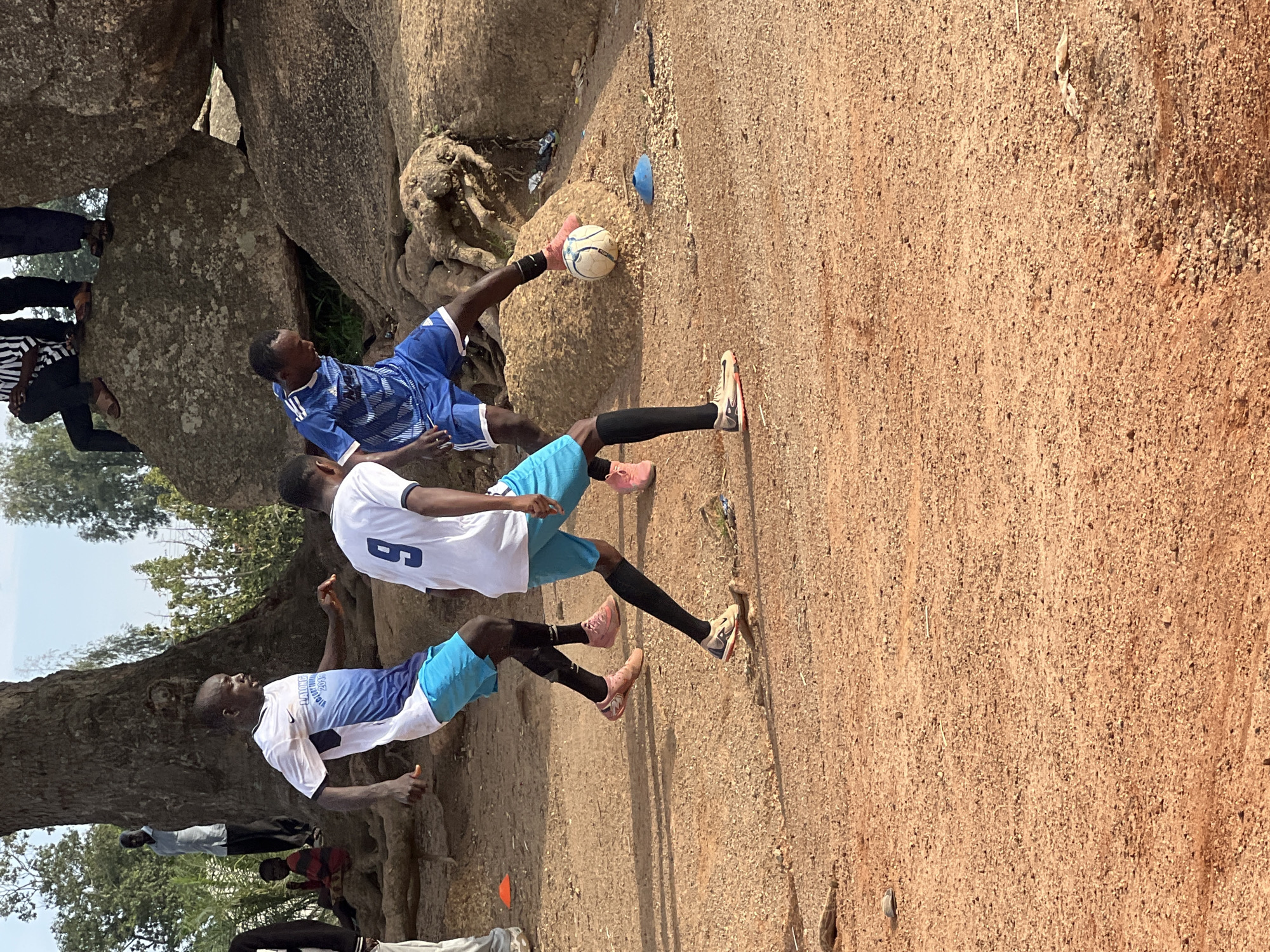 In Jos, Nigeria, three young men are engaged in a soccer game on a dirt field at the base of a large rock formation and a massive tree. One man in a white and blue jersey with the number "9" on his back is in the foreground, facing left and dribbling a white soccer ball with his right foot. He wears blue shorts, black socks, and pink soccer cleats. To his left, another man in a white and blue jersey and blue shorts is also playing, with his body angled towards the ball and his left foot extended. He is wearing pink soccer cleats and black socks. To the right of the man with the number "9", a third man in a blue and white striped soccer jersey and blue shorts is visible, also looking at the ball. He is wearing black socks and pink cleats. The ground is covered in reddish-brown dirt and small stones. In the background, the large rock formation is prominent, with some vegetation growing around its base. A large tree with exposed roots is to the left of the rocks. Other individuals are partially visible in the background, including a person standing near the tree and others sitting on the rocks. The sky is a pale blue.