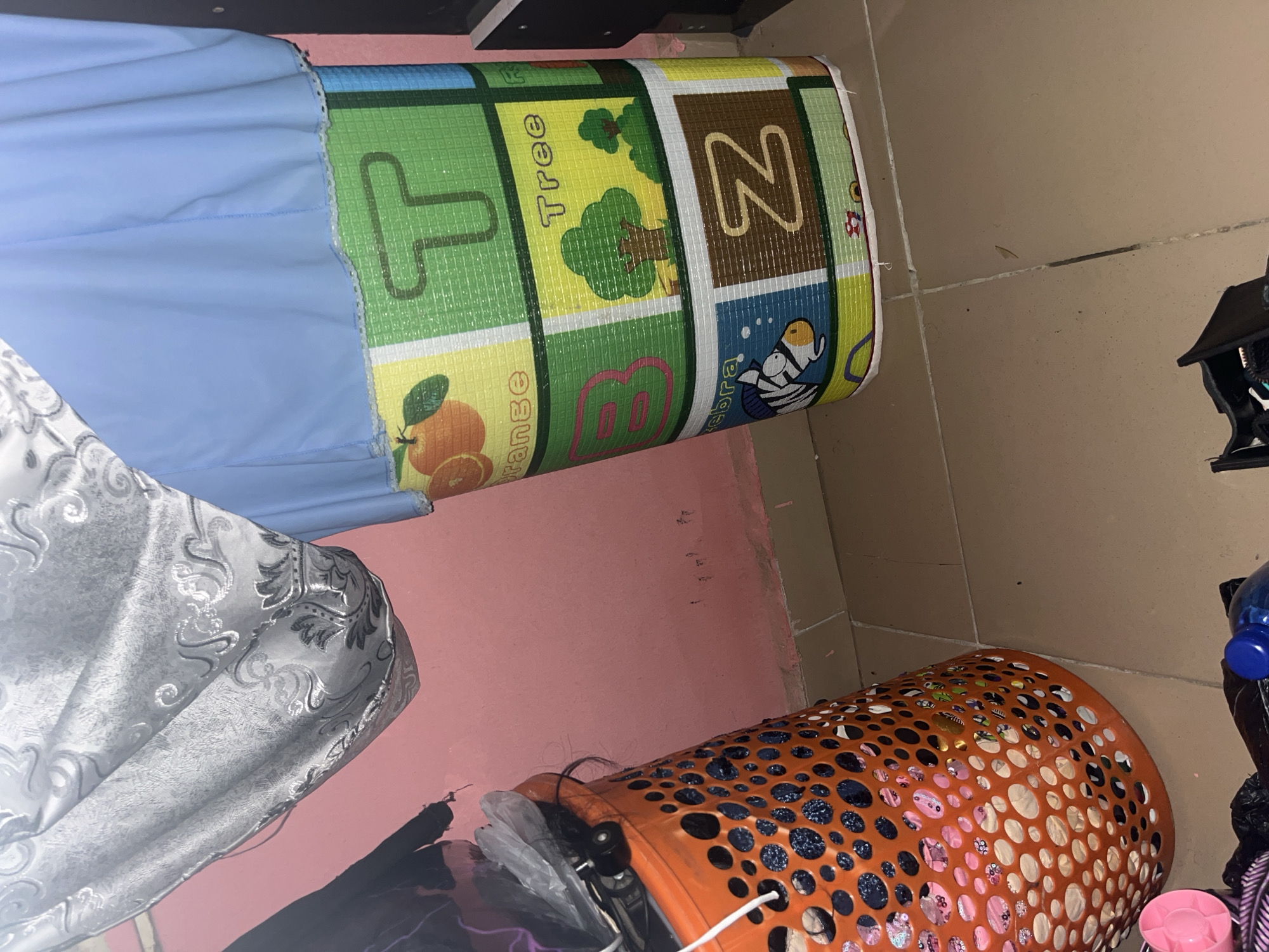 The image shows a close-up view of a room, possibly a bedroom or storage area, in Jauro Yinu, Nigeria. The wall is painted pink. In the foreground, an orange plastic laundry basket is filled with items. To the right of the basket is a rolled-up mat with colorful illustrations of letters of the alphabet and corresponding images, such as an orange for 'O', a tree for 'T', and a zebra for 'Z'. A blue cloth is draped over the top of the mat. In the lower part of the image, on the tiled floor, are some discarded items, including a pink object, a black plastic bag, and a blue bottle cap.