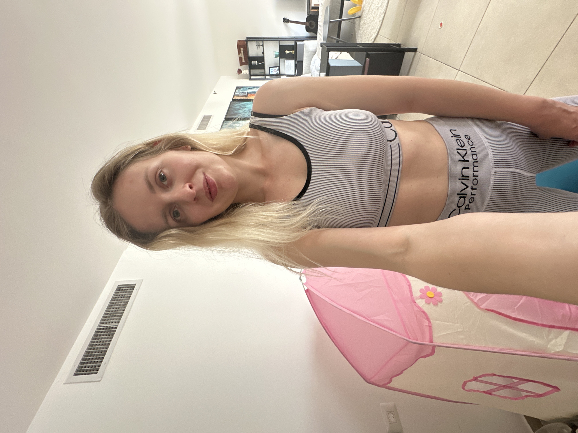 A woman is visible from the torso up, wearing a gray and white striped sports bra and matching leggings labeled "Calvin Klein Performance". She has blonde hair and light skin. The background shows a room interior. A white wall with a ventilation grill is on the left. A pink and white play tent is partially visible in the foreground. Further back, there is furniture including a black shelf and a light-colored rug. In the middle distance, there are pictures on the wall. Based on the objects and surroundings, this image appears to be taken in a domestic environment. There is no specific indication of Petah Tikva, Israel, in the image.