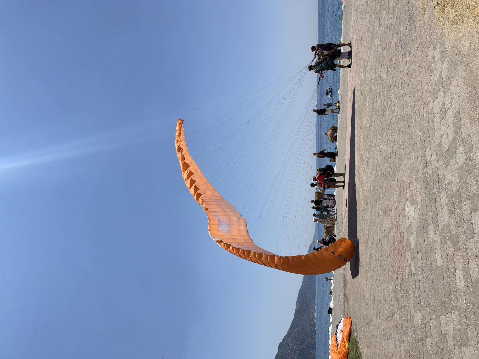 A clear, sunny day at a paragliding launch site on the coast in Fethiye, Turkey. The foreground features a paved walkway with interlocking rectangular stones, bordered by dry grass on the right. In the middle ground, a large, orange paraglider wing is spread open, its lines extending upwards towards the clear blue sky. Several people are gathered around, some appearing to prepare for a flight. One individual is seated in a paragliding harness, connected to the lines, while others stand nearby, holding the paraglider.
Further back, the turquoise water of the sea stretches to the horizon, with a distant, green, mountainous coastline on the left. A few beach umbrellas and some people are visible along the shoreline. The sky is a vibrant, cloudless blue, with a bright sun creating a lens flare effect. The overall atmosphere suggests an active and recreational environment, characteristic of a popular tourist destination.