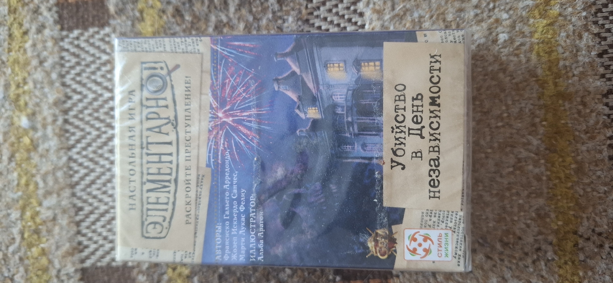 A vertically oriented, eye-level, slightly angled medium shot shows a board game box resting on a brown, textured fabric surface. The box is predominantly blue and depicts a dark, ominous house with lights on in the windows, set against a backdrop of fireworks. The title of the game, "Убийство в День Независимости" (Murder on Independence Day), is prominently displayed in a distressed font across the bottom half of the box, overlaying a tan-colored banner. Above the title, the game's name "ЭЛЕМЕНТАРНО!" (Elementary!) is written in a large, stylized font with a magnifying glass icon. Text indicating authors and illustrators is visible on the left side of the box. A small circular logo with abstract red and white figures is located in the lower-left corner of the box, next to the text "СТИЛЬ ЖИЗНИ" (Lifestyle). The fabric surface appears to be a rug or blanket with a repeating geometric pattern in shades of brown and yellow. There is no indication of the city of Tsimlyansk, Russia, in the image.