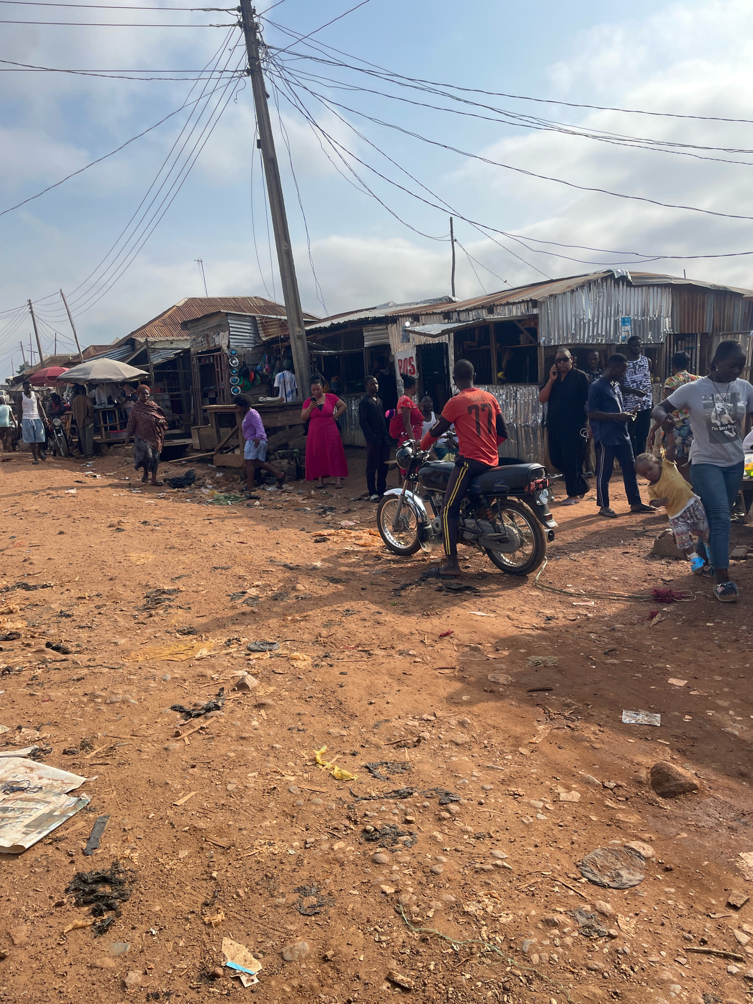 The image depicts a street scene in Kuruduma, Nigeria. The ground is composed of reddish-brown dirt, scattered with debris including plastic bags, wood fragments, and other refuse. Along the side of the road are numerous makeshift stalls constructed from corrugated metal and wood, indicating a market or commercial area. Overhead, a network of electrical wires crisscrosses the sky, supported by a concrete utility pole.
Several people are present in the scene. In the foreground, a young child in a yellow shirt and patterned shorts is being pulled by a woman in a grey t-shirt and blue jeans, who is holding the child's arm. Behind them, a man wearing an orange t-shirt with the number '77' is seated on a motorcycle. Other individuals are visible walking, standing, and interacting in the vicinity of the stalls. The stalls themselves appear to be selling various goods, though specific items are not clearly discernible. The overall impression is one of a bustling, informal marketplace in an urban setting.