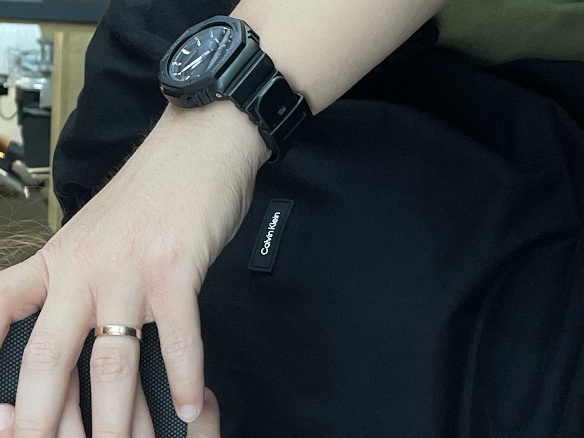 A close-up shot focuses on a person's left hand and arm, adorned with a rose gold ring and a black wristwatch. The hand rests on a black t-shirt with a "Calvin Klein" logo tag. The background is blurred, showing a glimpse of what appears to be a domestic setting with light-colored furniture and objects on shelves, possibly indicating an indoor environment within a home or apartment. There is no indication of Rostov-on-Don, Russia in this image.
