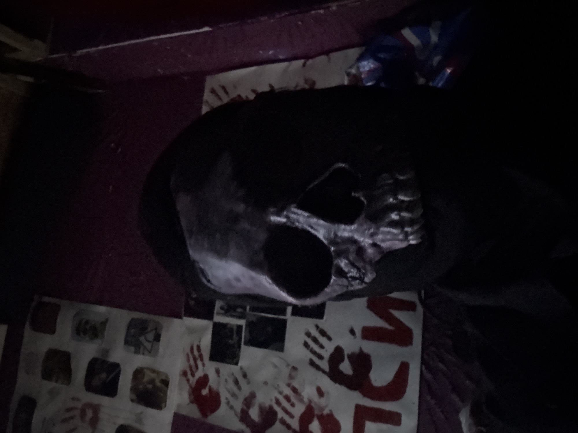 The image shows a silver skull mask with dark eye sockets and a black hood. Below the mask is a poster with images and bloody handprints. There is a piece of packaging behind the mask, and some text in red writing is partially visible below the poster. The background is dark and appears to be part of a room with a purple-colored wall or shelf. The location in the city of Saratov, Russia, is unknown.