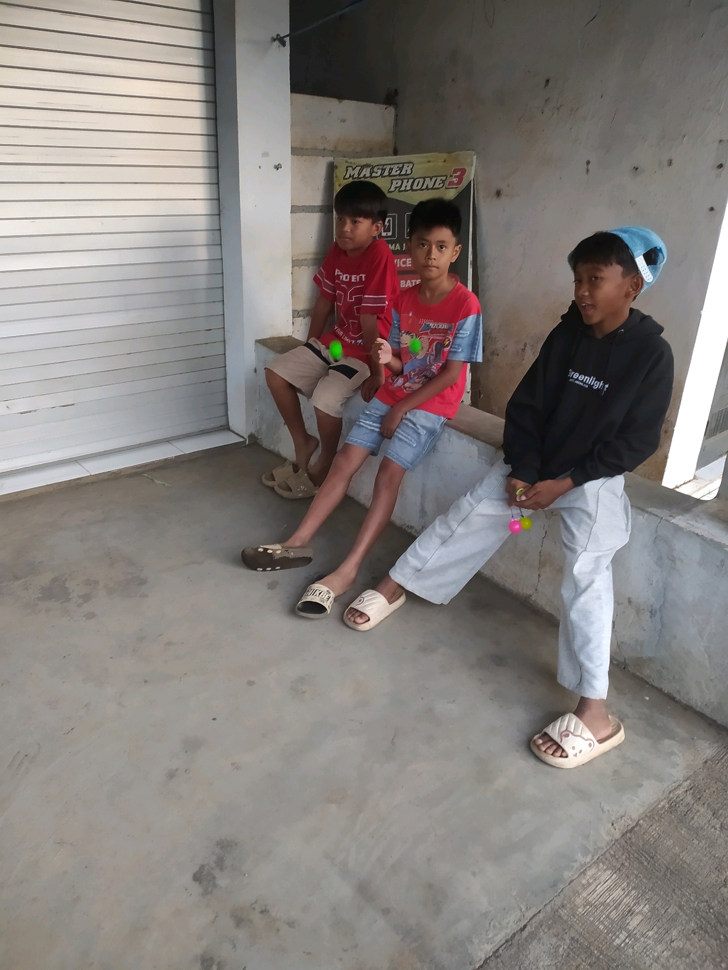 Three young boys are sitting on a concrete ledge outdoors, in front of a roll-up metal door. The boy on the left wears a red t-shirt and beige shorts. The middle boy is in a red t-shirt with blue sleeves and denim shorts. The boy on the right is wearing a black hoodie and light grey pants. He has a blue cap on his head. The middle and right boys are holding toys. The environment appears to be an urban setting in Bantaran, Indonesia, with a rough concrete floor and a textured wall behind them. The lighting suggests it is daytime.