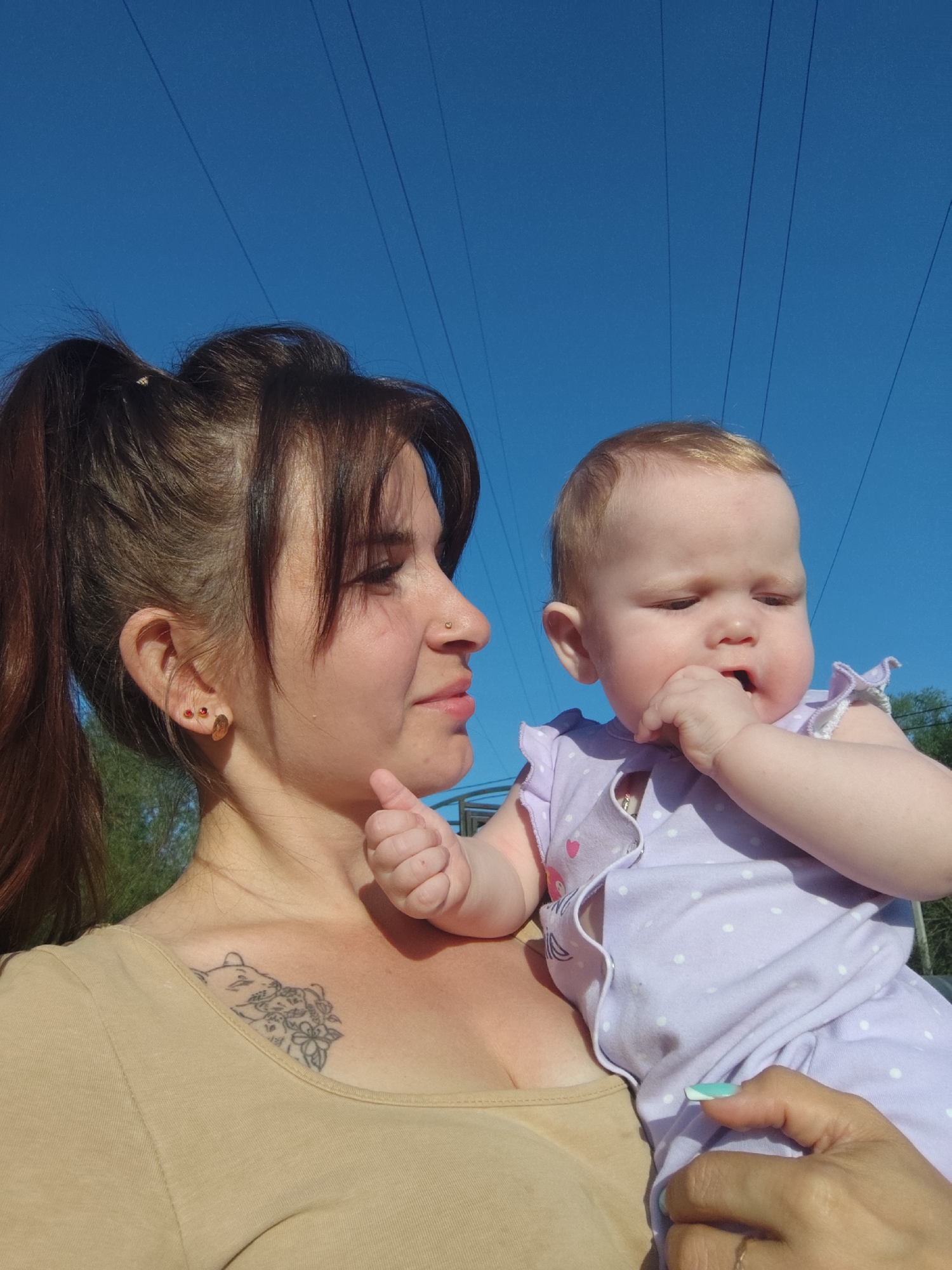 A woman is holding a baby. The woman has brown hair in a high ponytail, bangs, and a nose piercing. She is wearing a tan shirt and has a tattoo on her upper chest. The baby is wearing a lavender polka-dot outfit and has a finger in its mouth. The background shows a clear blue sky and power lines. The location is Фоки, Russia.