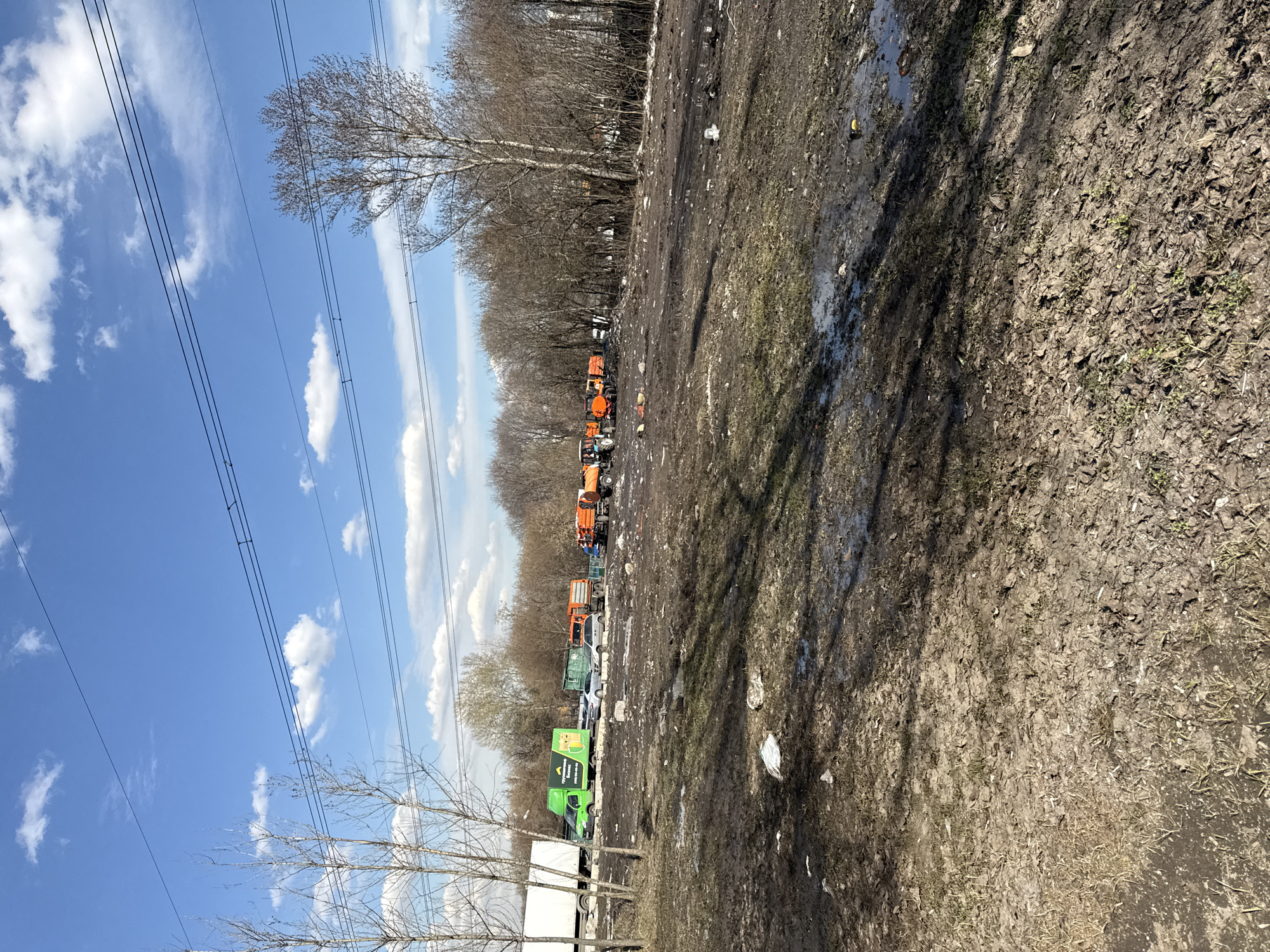 The image shows a muddy, debris-strewn area next to a line of parked trucks and vehicles. The vehicles are diverse, including what appear to be orange utility trucks, a white van, and a green truck with advertisement on it. A dirt surface covered with leaves, debris, and puddles occupies much of the foreground. A line of trees borders the vehicles, and a blue sky with scattered clouds is visible above, crossed by power lines.
Based on the presence of particular orange utility trucks and green trucks, as well as the general look of the landscape and street debris, it is possible that this scene is from Moscow, Russia. No people or other subjects are identified in the image.