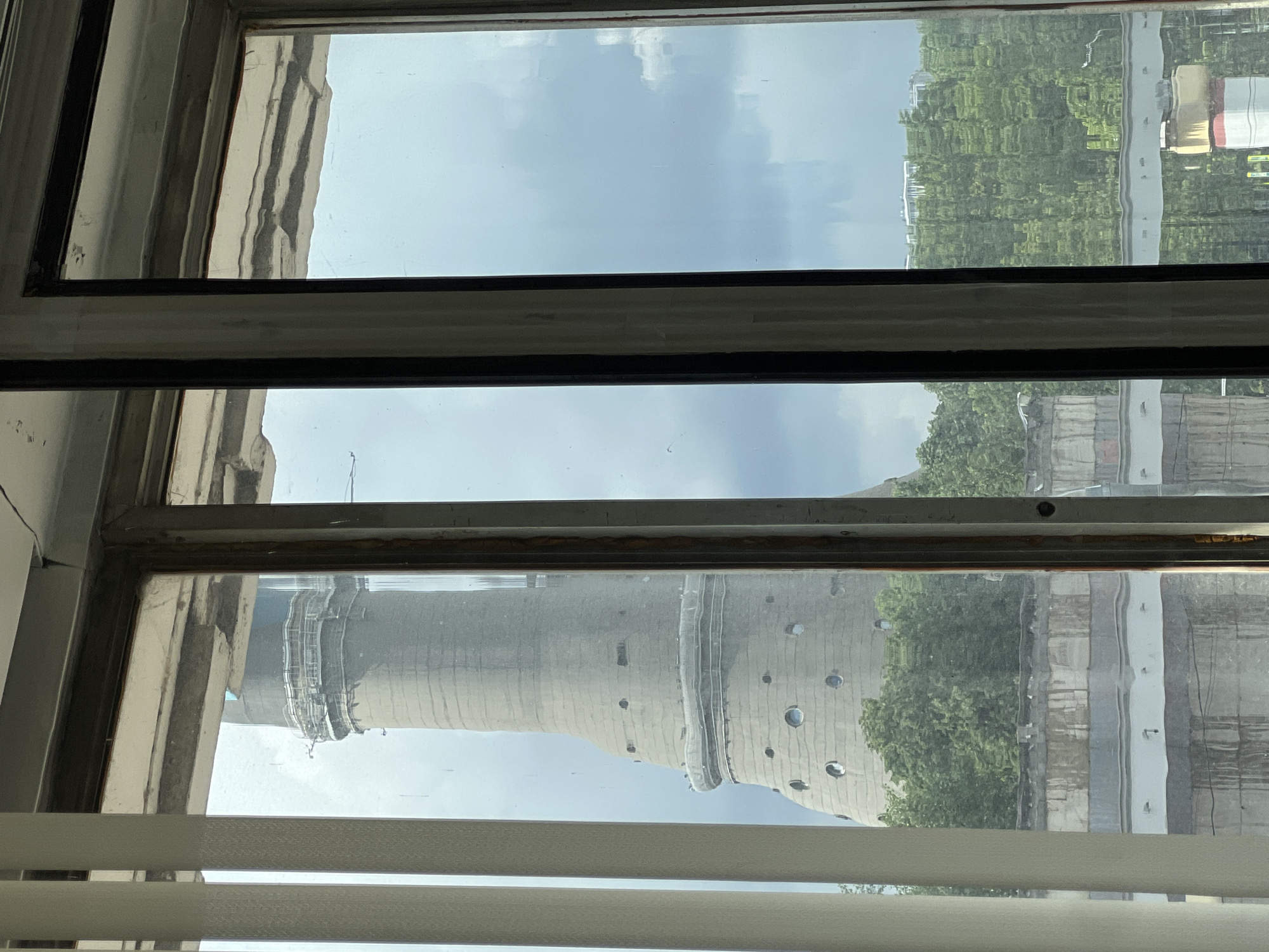 The image depicts a view through a window, focusing on the reflection of a structure outside. The reflection includes a tall, cylindrical tower with a visible pattern of circular windows and horizontal banding. This suggests the building is of industrial or utility design. The windows are reflecting the tower and some green foliage and buildings. The window frame itself is aged, with visible wear and discoloration. Blinds partially obscure the lower portion of the window. Considering the location is Khimki, Russia, the tower likely corresponds to the Moscow State University of Culture and Arts building.