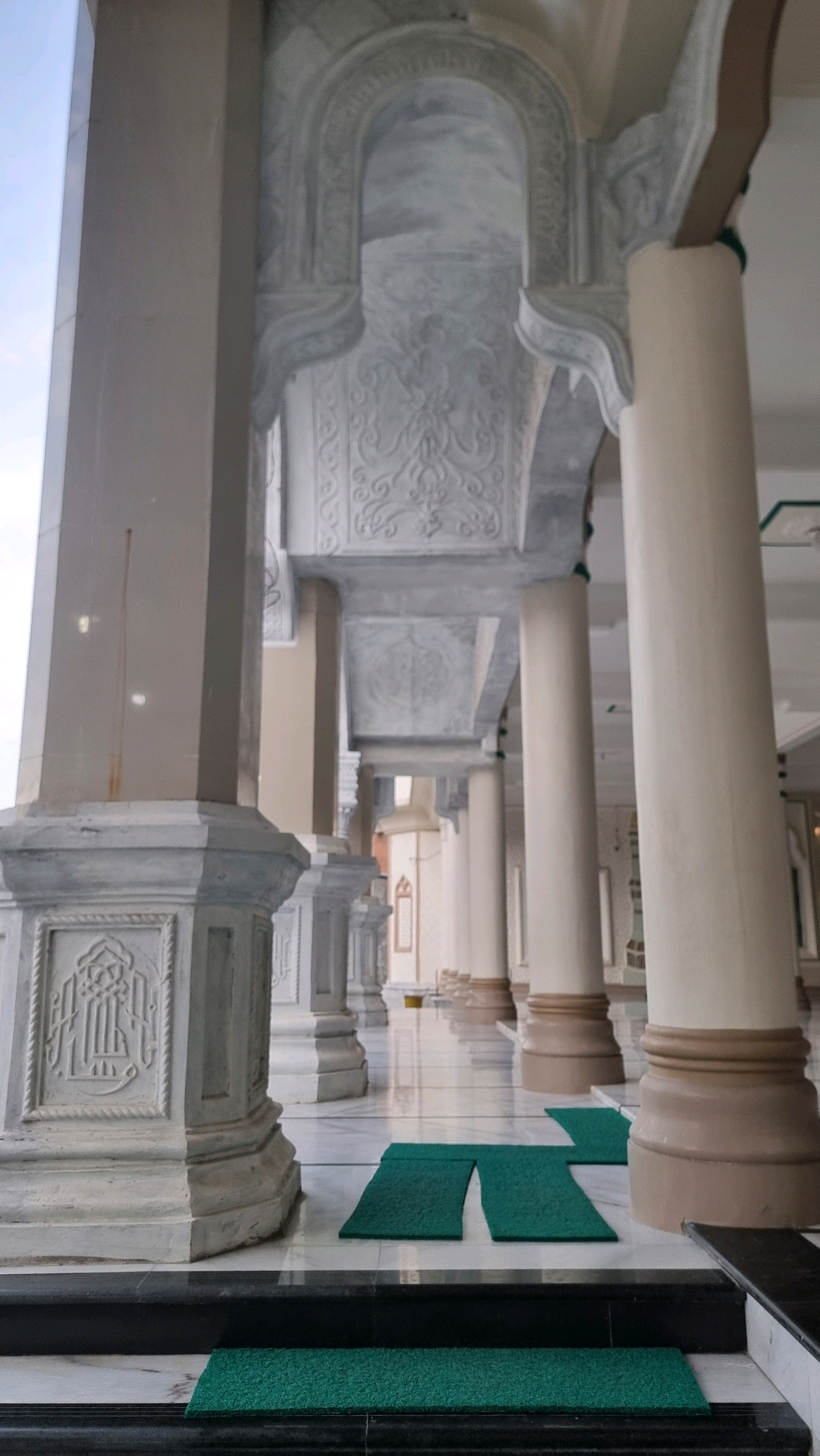 The image shows a colonnade of a mosque in Lhokseumawe, Indonesia. The architecture features large, cream-colored columns with brown bases and intricate white decorative carvings on the upper sections and the ceiling. The floor is polished marble with a black and white tiled border. Several small, green prayer mats are placed on the floor. In the background, more columns and arched structures are visible, suggesting a long, covered walkway or prayer area. The overall impression is one of grandeur and solemnity.