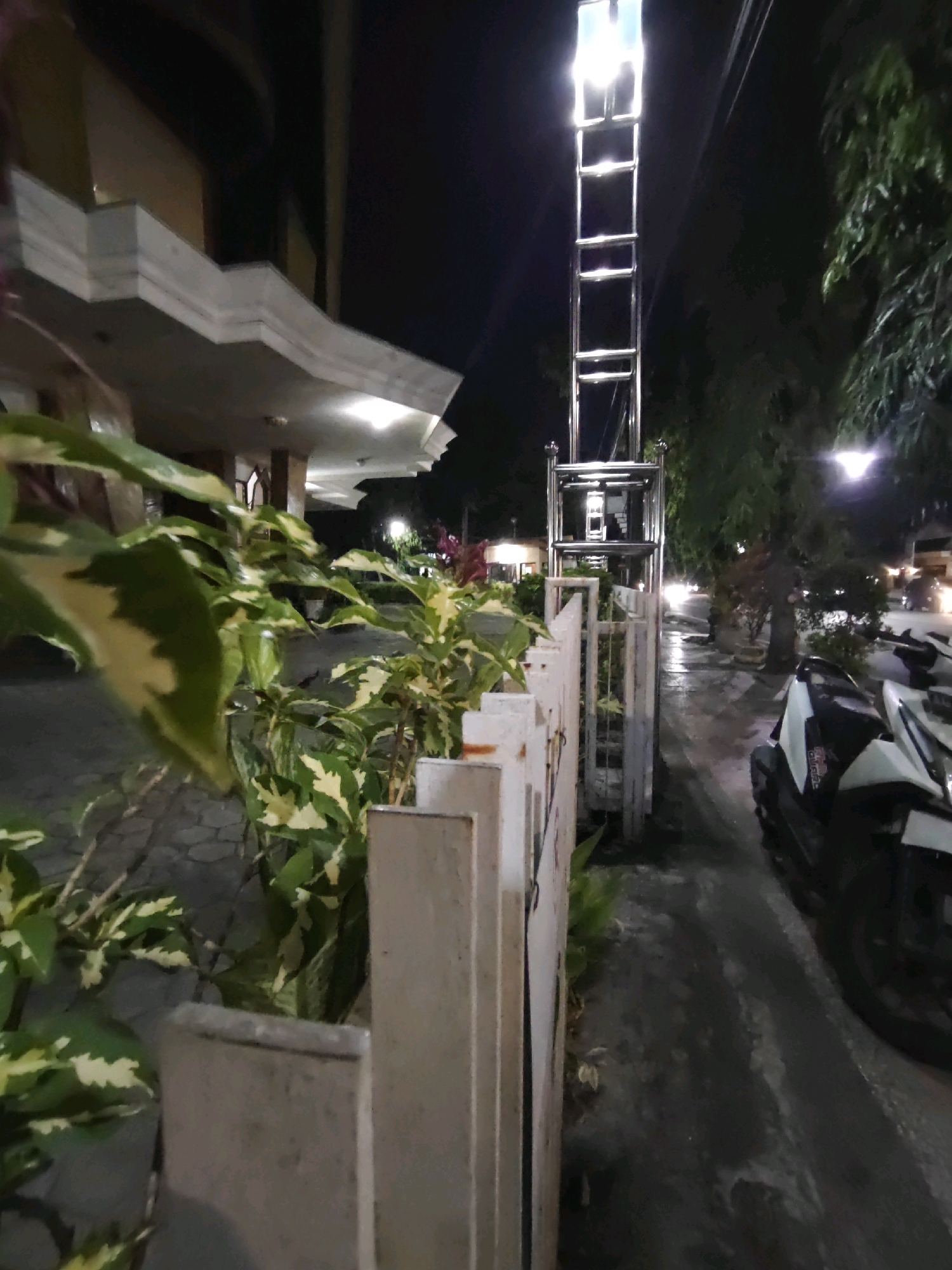 The image captures a nighttime street scene in Kediri City, Indonesia. In the foreground, there are lush green plants with variegated leaves and a low white fence. To the right, a white scooter is parked on a wet asphalt road, suggesting recent rain. Further down the street, a tall, multi-level structure with a bright light at the top stands, possibly an elevator or a promotional display. Beyond that, a building with a modern architectural overhang is visible. The scene is illuminated by streetlights, the light from the tall structure, and the headlights of vehicles in the distance. The overall impression is one of a well-lit urban environment at night.