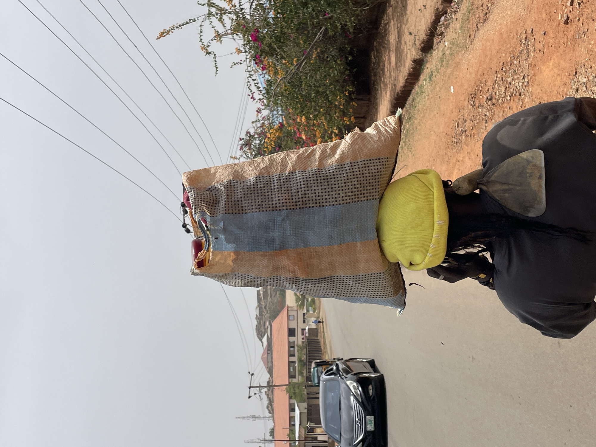 A man sits on a motorcycle on a paved street. He is wearing dark clothing and carrying a shovel. Behind him are two large bags, one striped and the other yellow. The motorcycle is stopped on a street with several vehicles in the background, including a black car. Buildings and vegetation are visible along the street, and power lines stretch across the sky. The scene is located in Jos, Nigeria.