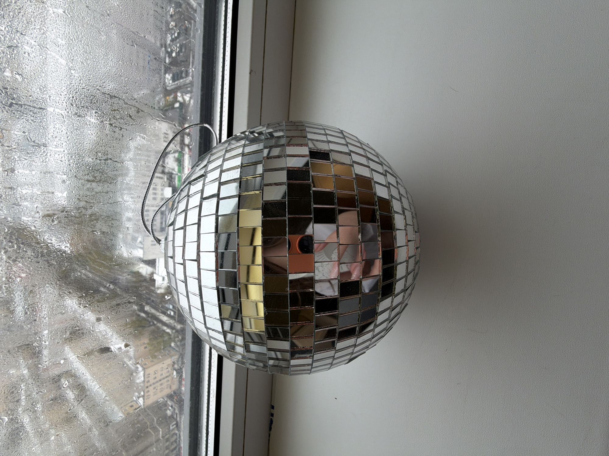 The image displays a disco ball placed indoors next to a window, providing a view of an urban environment. The disco ball, a prominent spherical object, is covered entirely with small, square mirror tiles that reflect its surroundings. A metallic loop for hanging is visible at the top of the ball.
The windowpane on the left side of the image is heavily covered in water droplets or condensation, blurring the outdoor scene into indistinct shapes. Through the wet glass, blurred outlines of city buildings can be seen, indicating an urban setting and a high vantage point, possibly in Moscow, Russia as per the context. The condition of the window suggests rainy weather or high humidity outside.
To the right of the disco ball and the window frame, a plain, light-colored wall or window sill completes the composition.
A notable detail is the reflection captured within the disco ball's mirror tiles. In the lower-central part of the ball, a clear reflection of the person taking the photograph is visible, including a hand holding a smartphone (with an orange element, possibly a case) and a partial view of their face and upper body.
No specific activity or event is actively taking place; the scene is a static arrangement. The diffused light suggests it is daytime, possibly overcast. There is no visible text in the image.