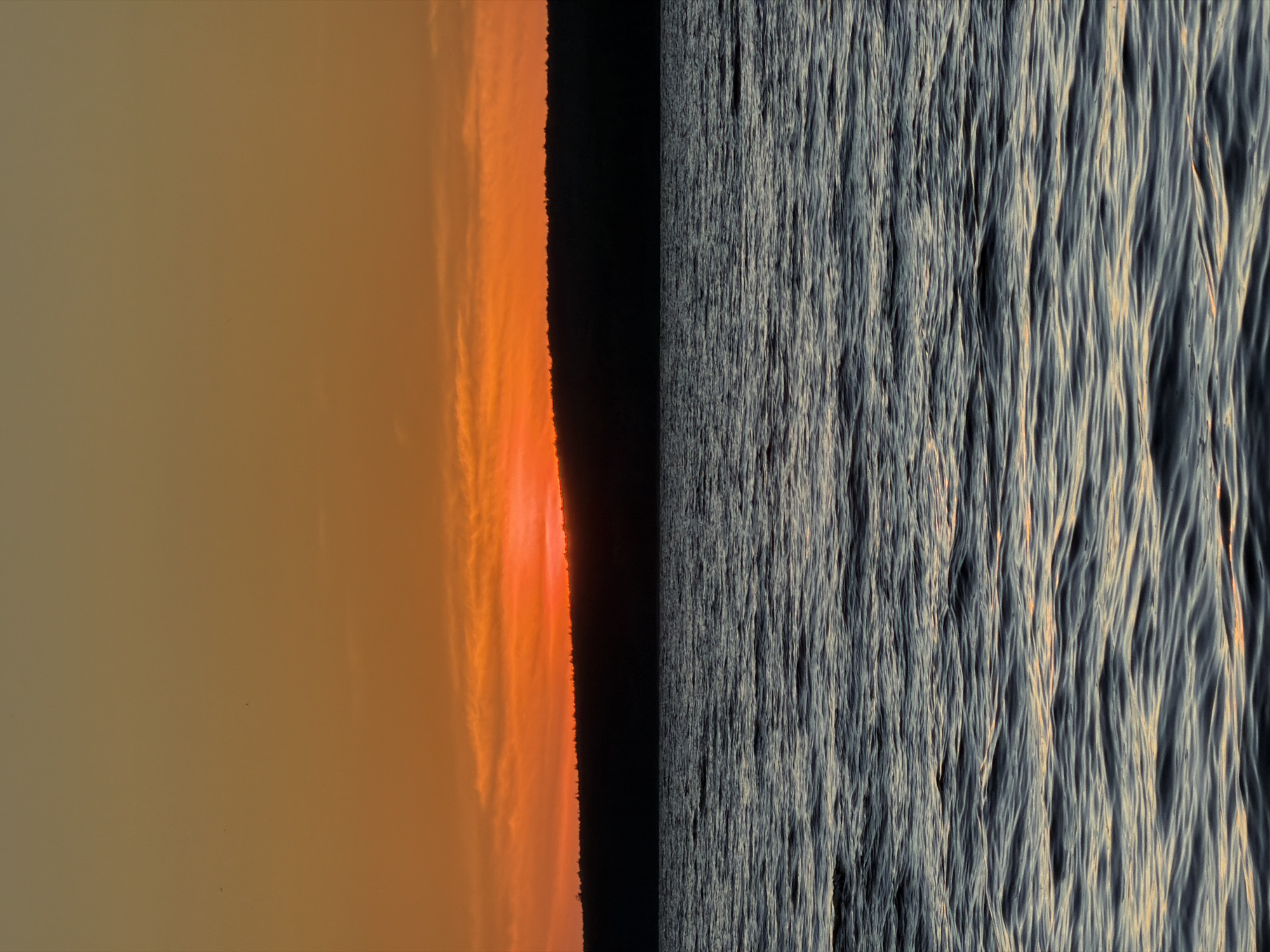 This is a photograph of a sunset over a body of water, likely a river or lake, with a tree-lined silhouette of a distant shore. The sky transitions from a pale orange at the top to a vibrant, fiery orange near the horizon, with thin, wispy clouds catching the last light. The water in the foreground is dark and rippled, reflecting the colors of the sky in muted tones. A dark, horizontal band of trees forms the silhouette of the land against the bright sky. There are no people, animals, or man-made structures visible in the image. The scene evokes a sense of natural beauty and tranquility. Without additional information or context, it is impossible to determine the specific location in Chaikovsky, Russia.