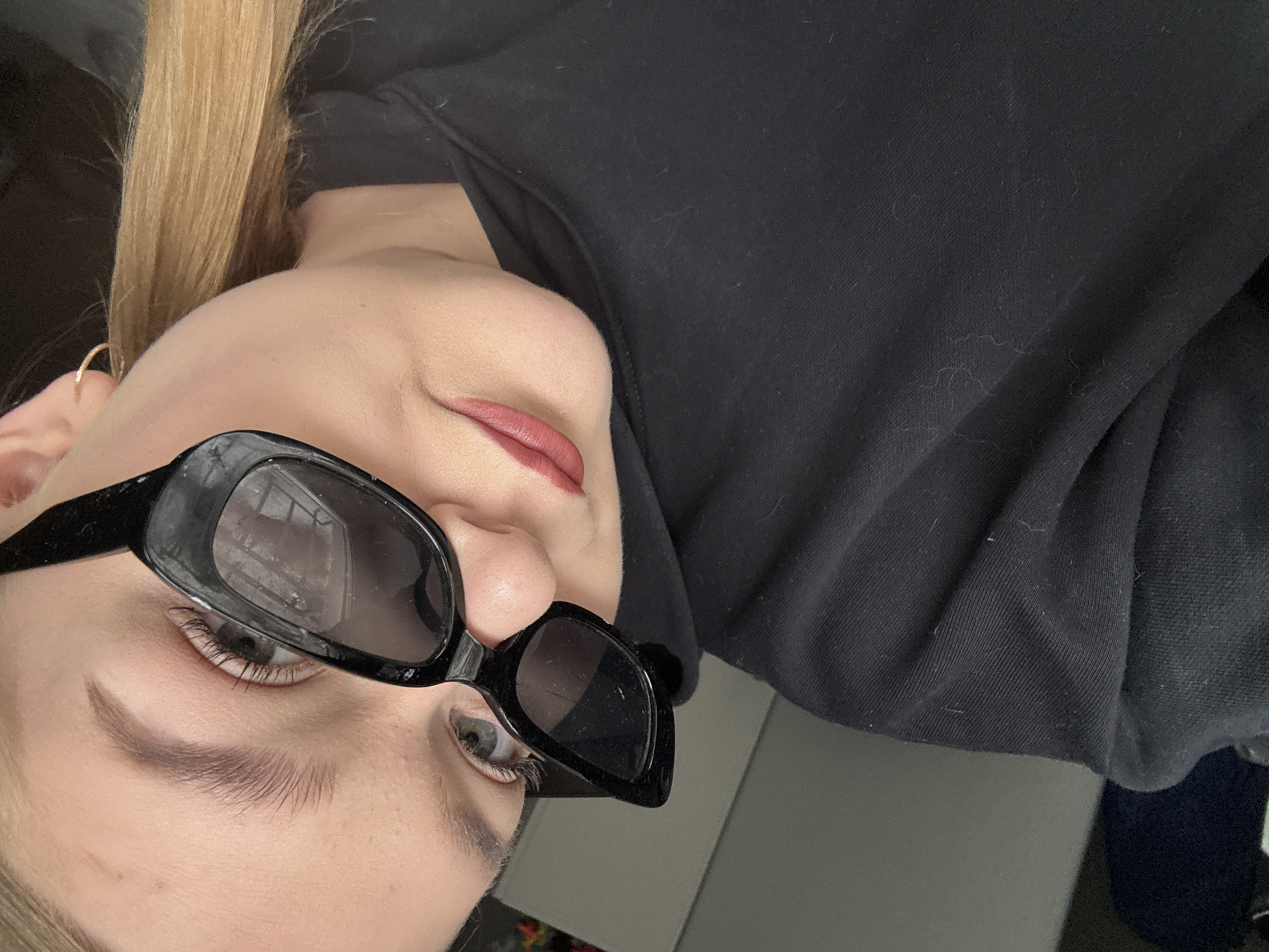 A selfie-style image of a woman with light blonde hair, wearing black, rectangular sunglasses and a dark-colored hoodie. She has fair skin, light eyebrows, and is wearing lip color. A gold hoop earring is visible in her left ear. The background is out of focus, but a gray-toned surface and dark objects can be discerned. The location is not identifiable as Chaikovsky, Russia from the provided image.