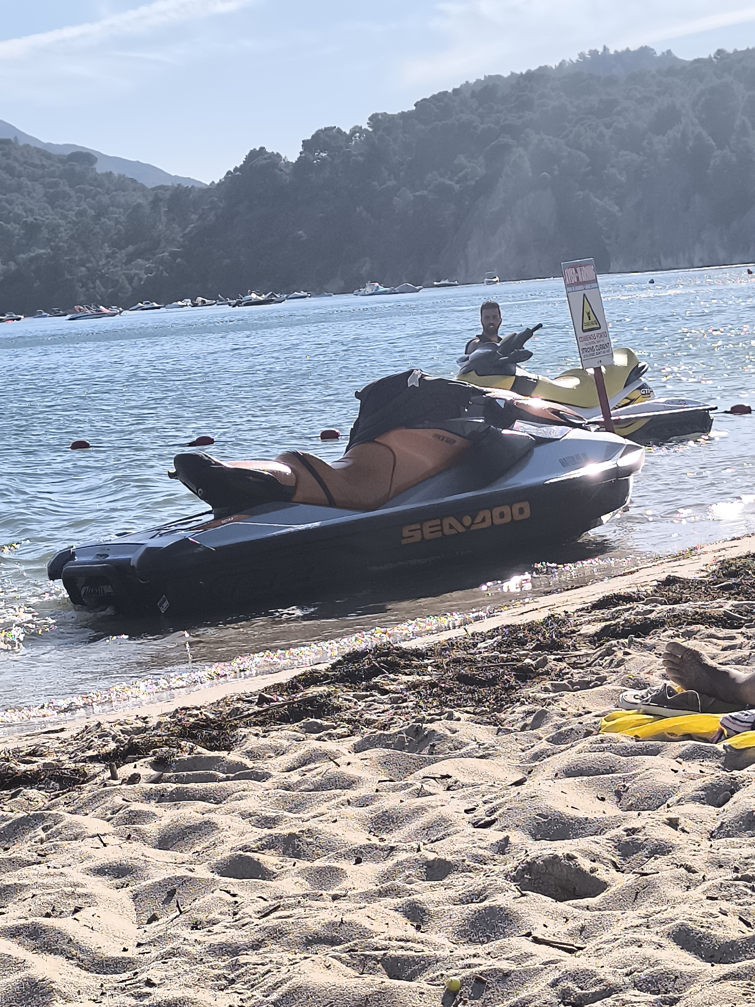 The image depicts a sunny day at a beach with calm blue water. In the foreground, two jet skis are partially submerged at the water's edge on a sandy shore littered with seaweed and driftwood. The jet ski closest to the viewer is black and grey with orange accents and the branding "SEA-DOO" is clearly visible. Behind it, a yellow jet ski is also in the water, with a person sitting on it. The person, a man with dark hair and a beard, is looking towards the camera.
In the background, the water stretches out, dotted with several other jet skis and a few buoys. Beyond the water, a lush green, tree-covered hill rises, with a clear blue sky above. The sun is casting bright reflections on the water. A warning sign is visible near the yellow jet ski, with text that appears to read "CAUTION FOR STRONG CURRENT".
On the sandy beach in the foreground, a person's feet and legs are visible, casually resting. Yellow flippers are lying next to them. The overall impression is a relaxed day at the beach, with water sports being enjoyed. The time of day appears to be mid-morning or early afternoon, judging by the bright sunlight and clear sky. The location is a beach, likely in a coastal area like Setúbal, Portugal, given the presence of hills and the general Mediterranean feel.