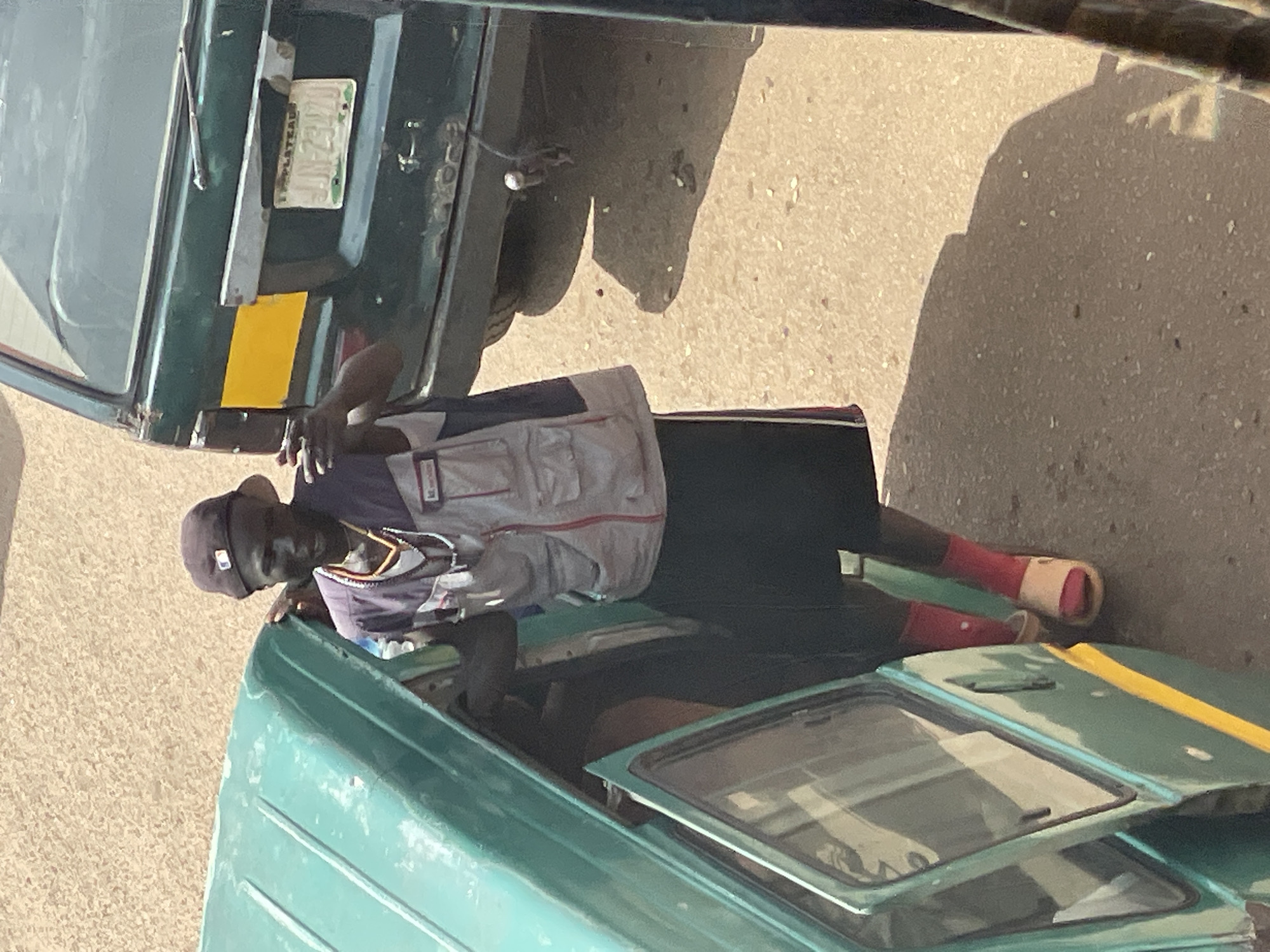 In this overhead shot, a dark-skinned person wearing a dark baseball cap, a grey vest over a purple shirt, black shorts, red socks, and sandals is standing next to the open door of a teal van. They are holding a mobile phone in their right hand. To the right of the van is a green vehicle with a yellow stripe on its side and a license plate that reads "JJN 23470". The ground is a sandy, light-brown color with scattered debris. The scene appears to be an outdoor urban environment.
