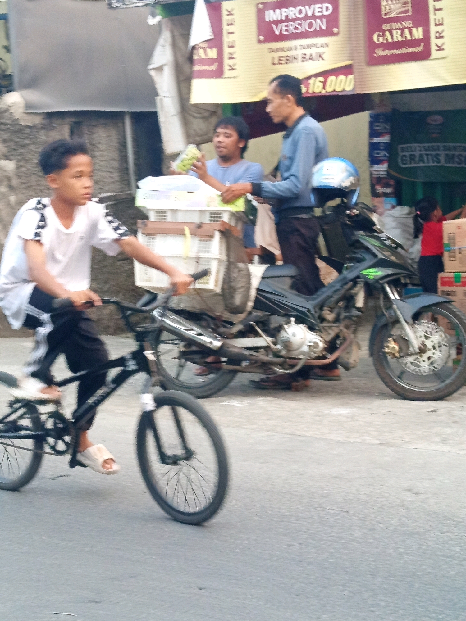 The image depicts a street scene in Karawang, Indonesia, with a young boy riding a bicycle in the foreground. The boy, wearing a white t-shirt with black stripes on the sleeves and black pants, is on a black bicycle with a silver exhaust pipe. He is wearing light-colored sandals. Behind him, two men are visible. One man, wearing a blue shirt, is holding a green bottle and appears to be interacting with the other man, who is partially obscured. A motorcycle is parked to the right of the men, with a blue helmet on its handlebars. In the background, there are structures with signage, including one that reads "GUDANG GARAM INTERNATIONAL" and another with "IMPROVED VERSION". The environment appears to be an outdoor urban setting, with a textured concrete ground. The lighting suggests it is daytime.