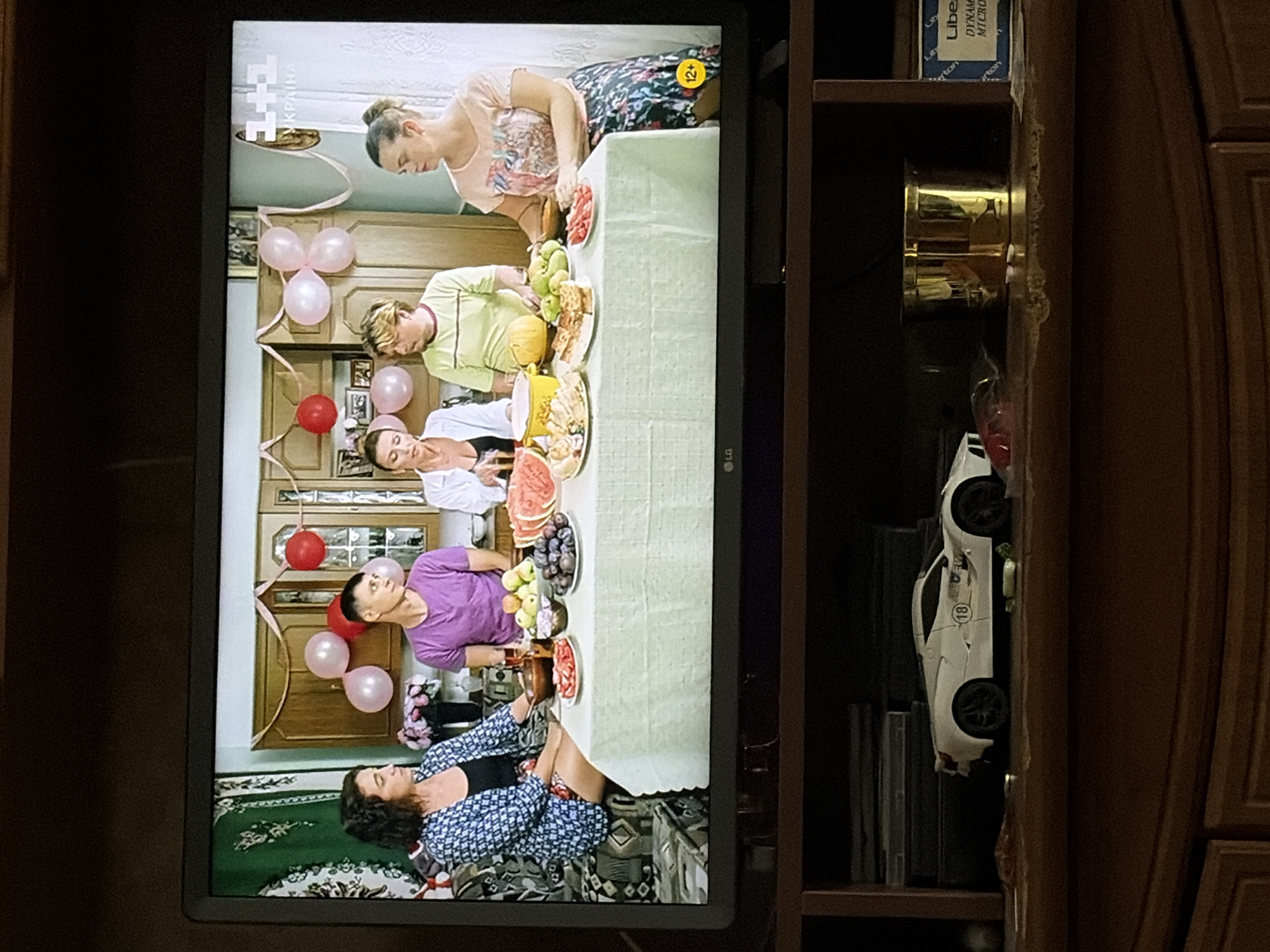 The image displays a television screen showing a scene with six people seated around a table laden with food. The people are positioned in what appears to be a domestic setting, possibly a dining room or kitchen, indicated by the cabinets and decor in the background. Balloons are visible, suggesting a celebration. The people are dressed casually. The television is placed on a wooden cabinet, and below the television, on the cabinet, a white toy car is visible. The overall lighting suggests indoor illumination. The location is unknown.